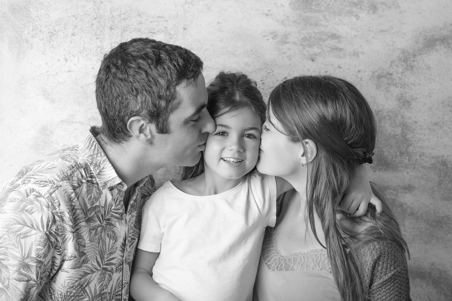 Black and white photo of parents kissing their smiling young daughter on both cheeks against a wall.