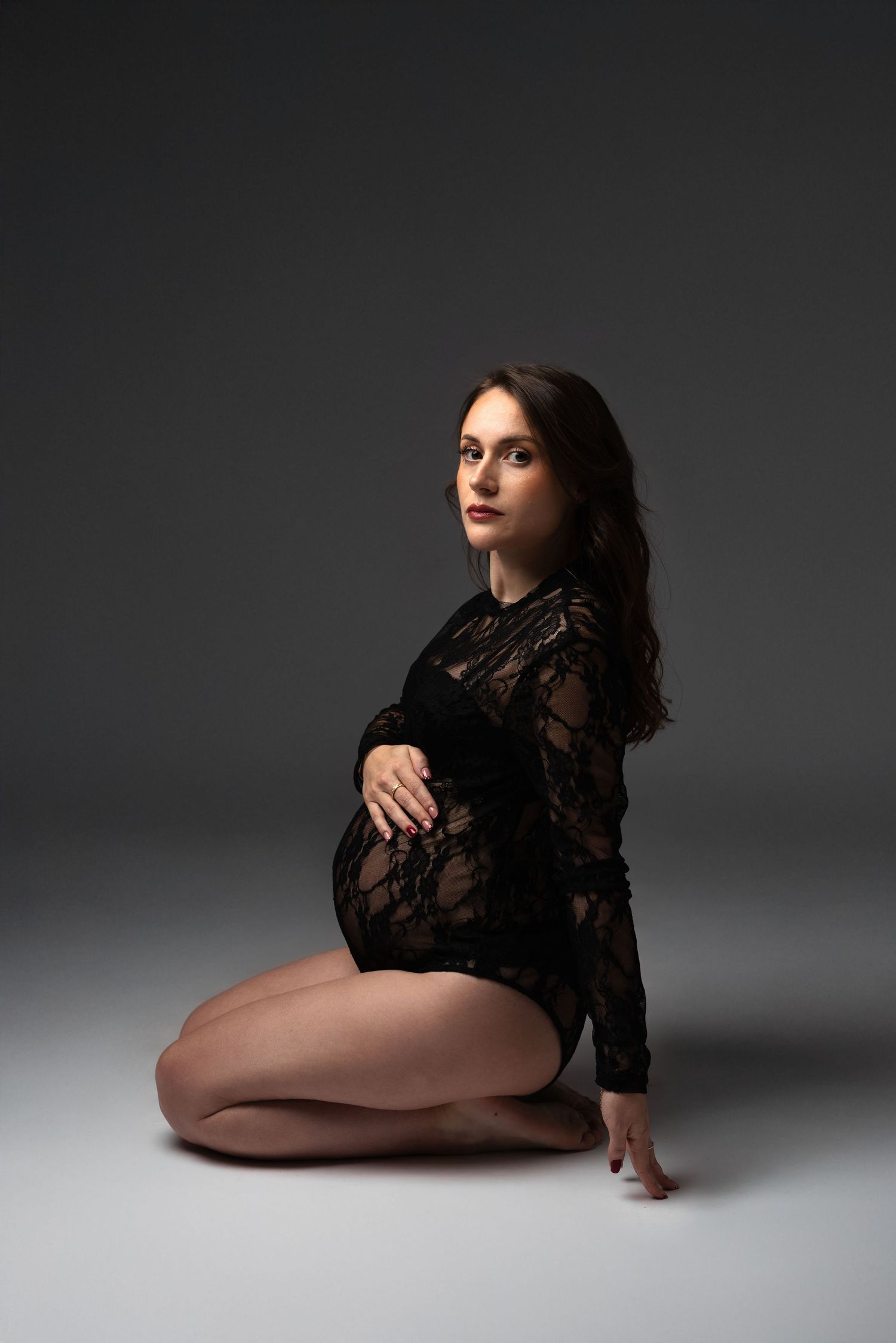 Pregnant woman in black lace bodysuit kneeling on studio floor, cradling baby bump, elegant maternity portrait.