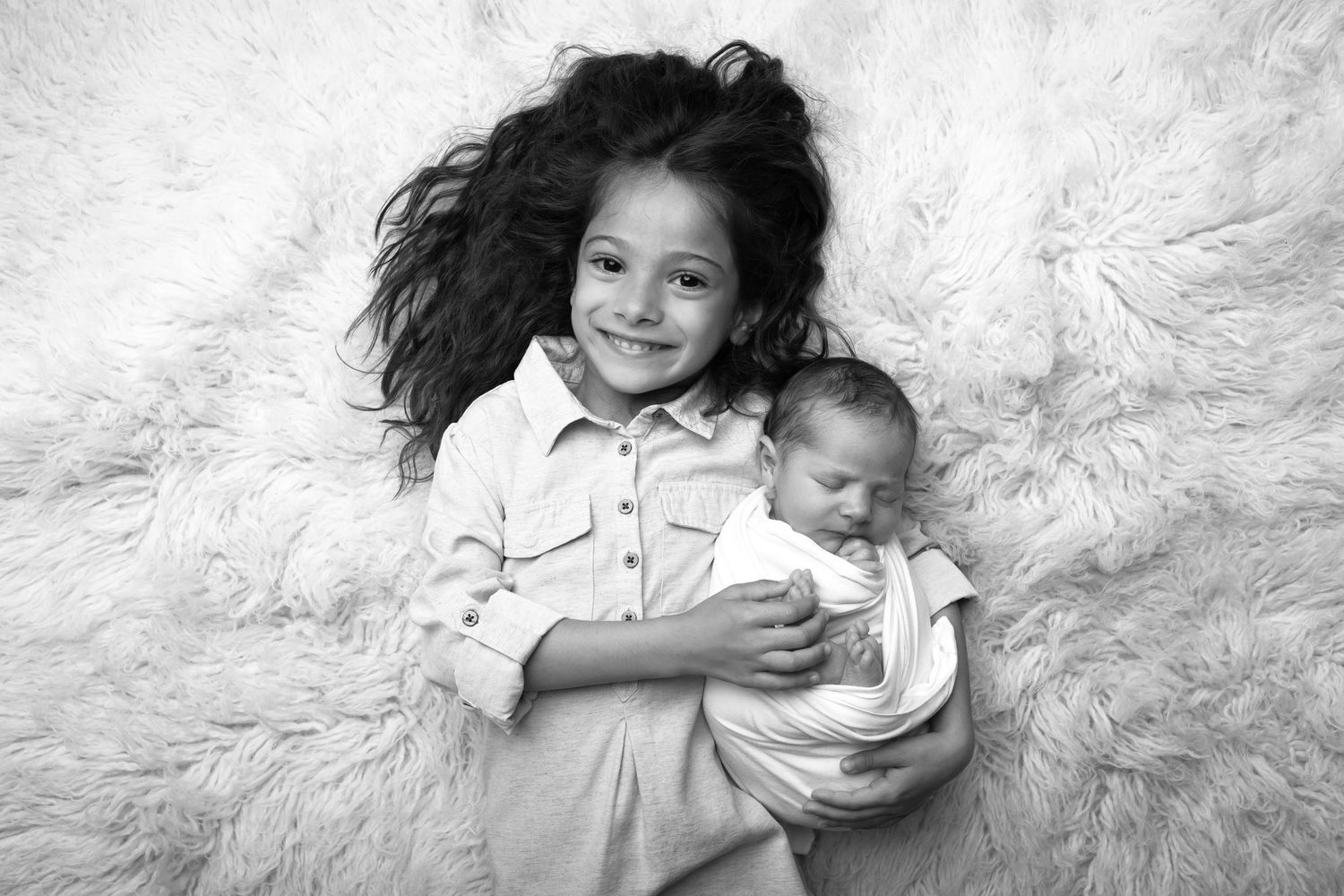 Black and white photo of a smiling girl with curly hair holding a swaddled newborn baby on a fluffy white rug.