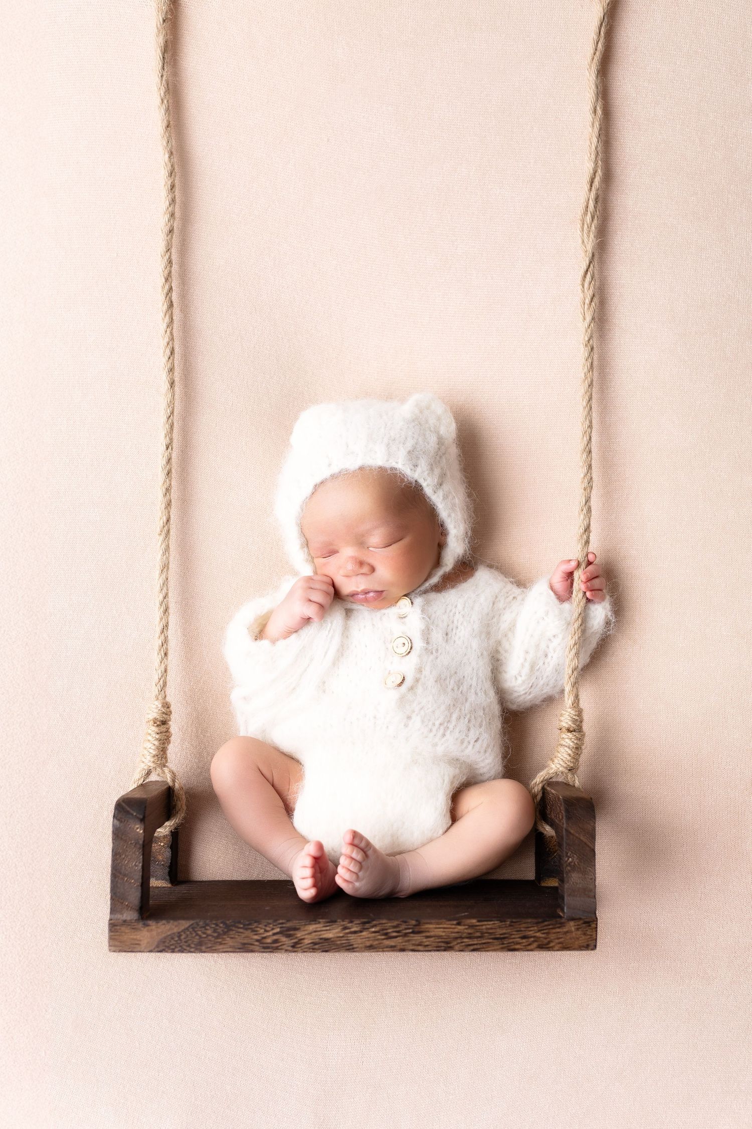 Peaceful newborn baby dressed in white fuzzy bear romper sleeping on a tiny wooden swing. séance naissance chavanoz lyon