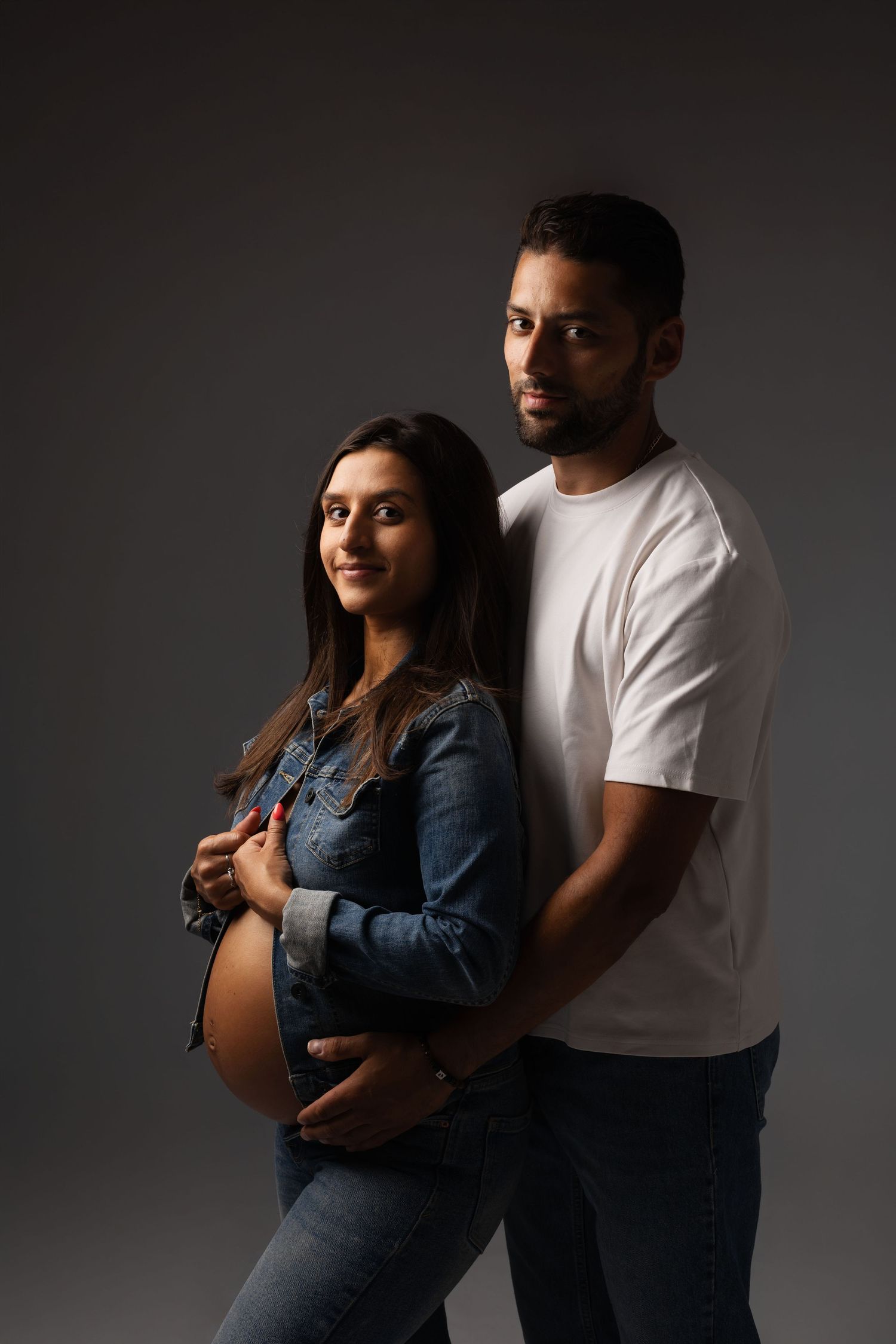 Pregnant woman in denim jacket with partner in white t-shirt posing together on gray background.