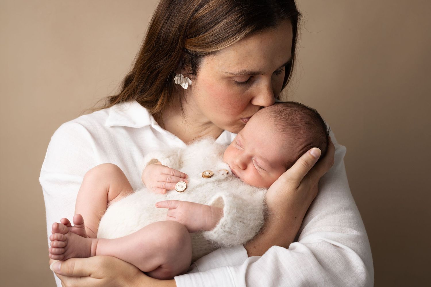 A mother in a white shirt tenderly kisses her sleeping newborn baby, held gently in her arms.