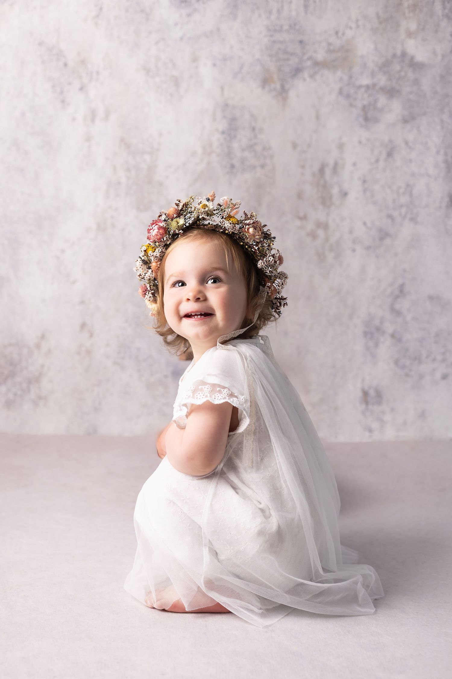A smiling baby girl in a white dress and flower crown kneels against a soft gray textured background.