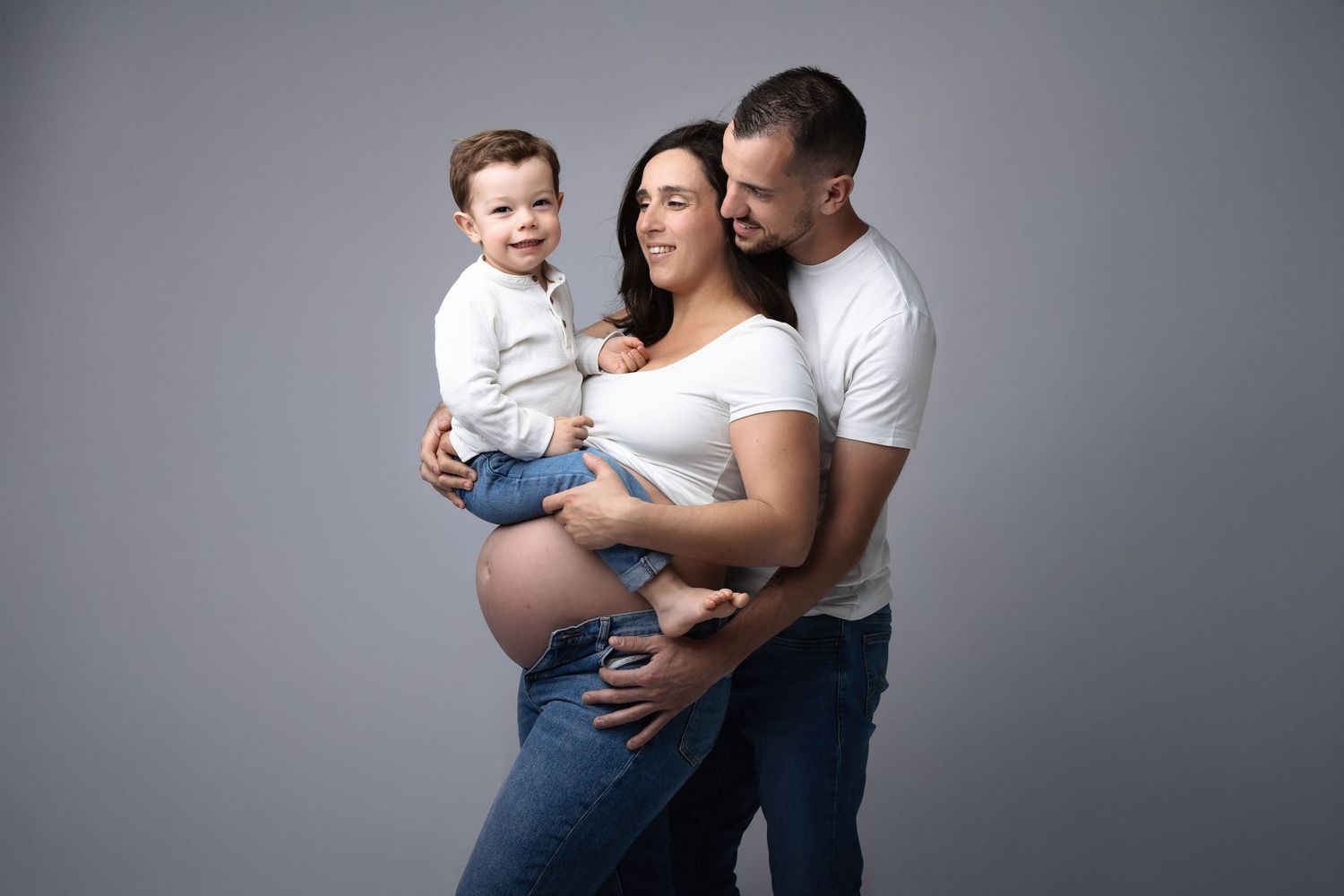 Smiling pregnant woman with husband and toddler son posing together on gray background. photographe grossesse chavanoz lyon