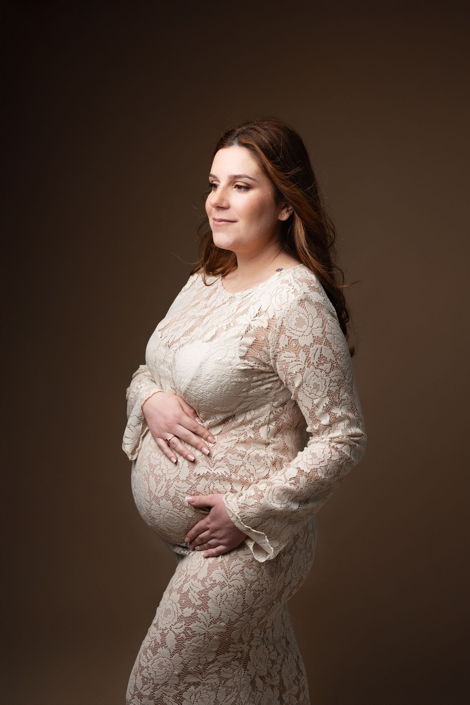 Smiling pregnant woman in white lace dress cradling her baby bump in a studio portrait. photographe grossesse chavanoz lyon