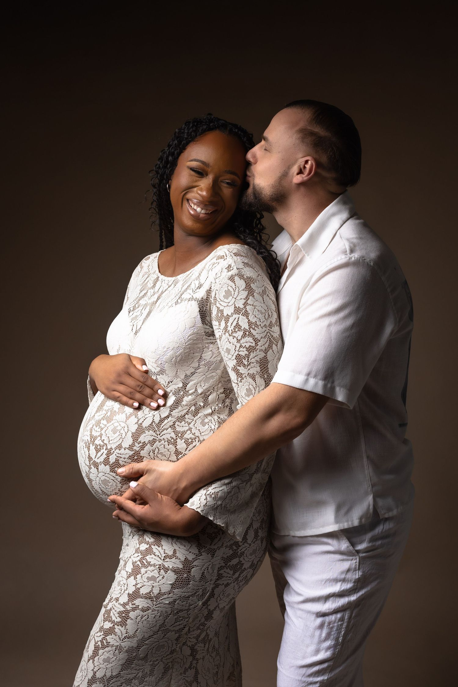 Smiling pregnant Black woman in white lace gown embraced by partner in white, studio maternity photo. photographe grossesse chavanoz lyon
