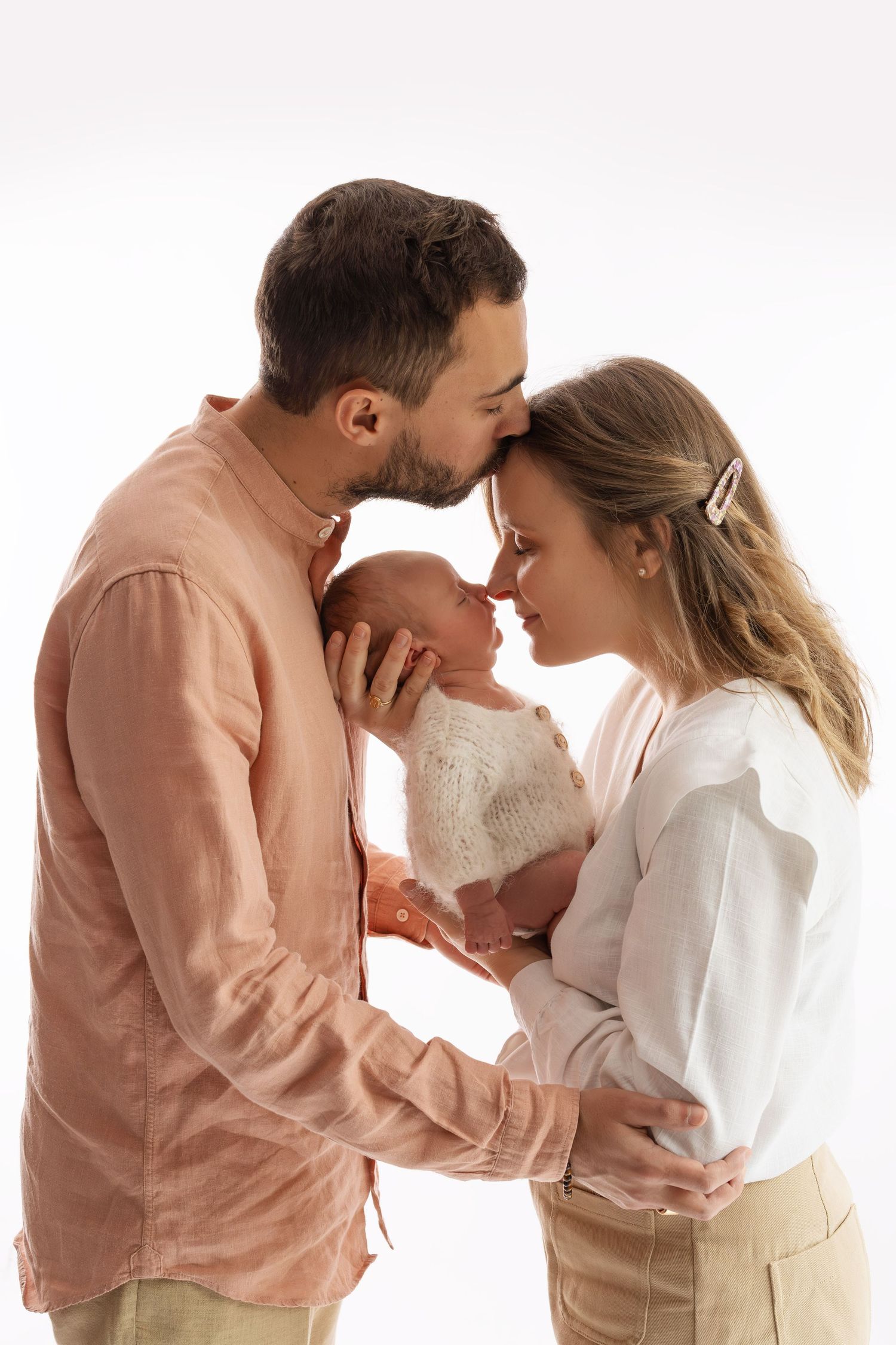 A couple tenderly holds their newborn baby, touching foreheads in a loving embrace against a white background.
