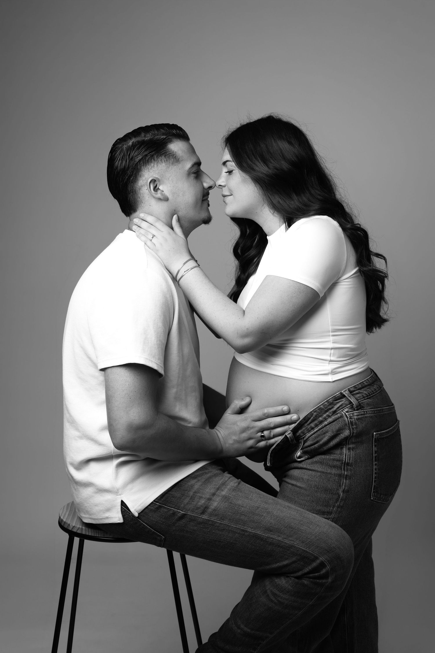 Black and white photo of a pregnant couple sharing a tender moment, man seated on stool embracing woman's baby bump.