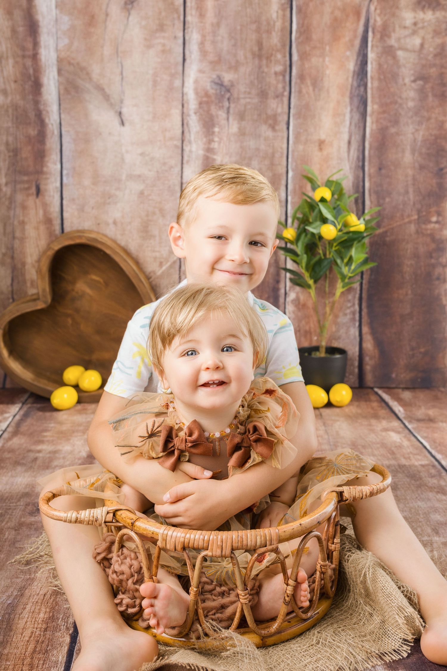 Two smiling blonde siblings pose together in a wicker basket with a rustic wood backdrop and lemon tree.