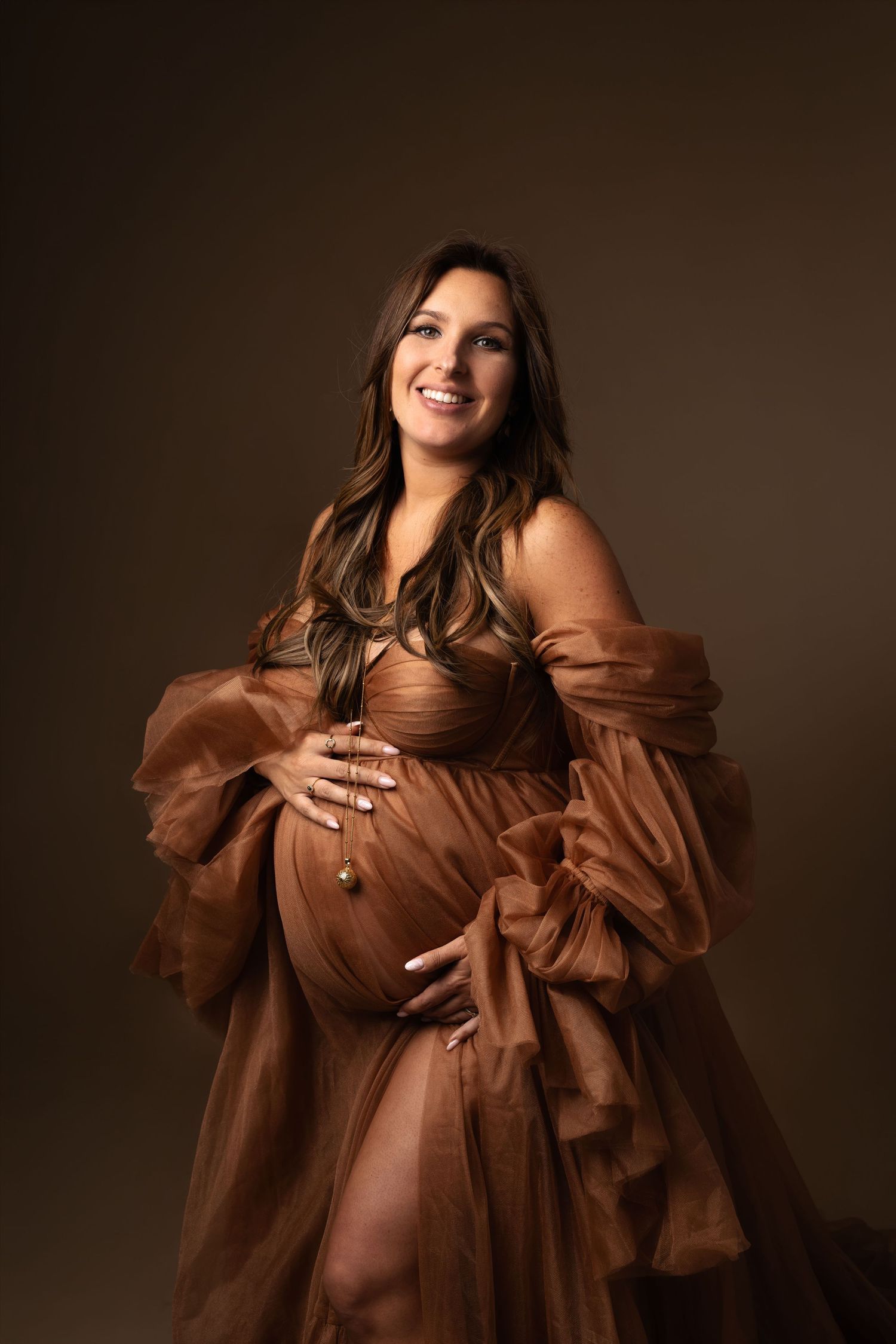 Pregnant woman in flowing brown tulle maternity gown cradling baby bump, smiling in studio portrait.