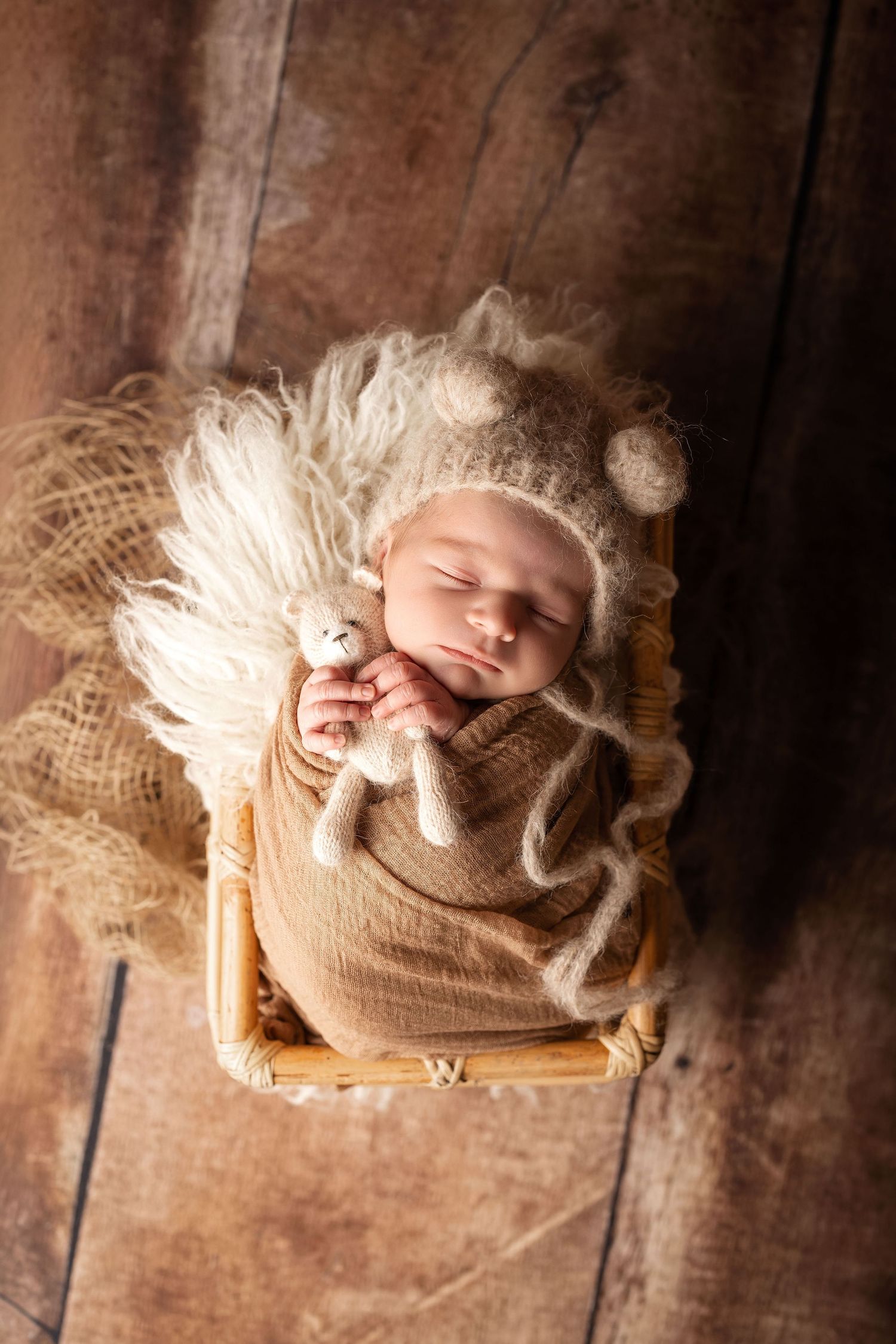 Sleeping newborn baby wrapped in beige swaddle holding tiny knit bear, resting in wooden crib with fluffy blanket.