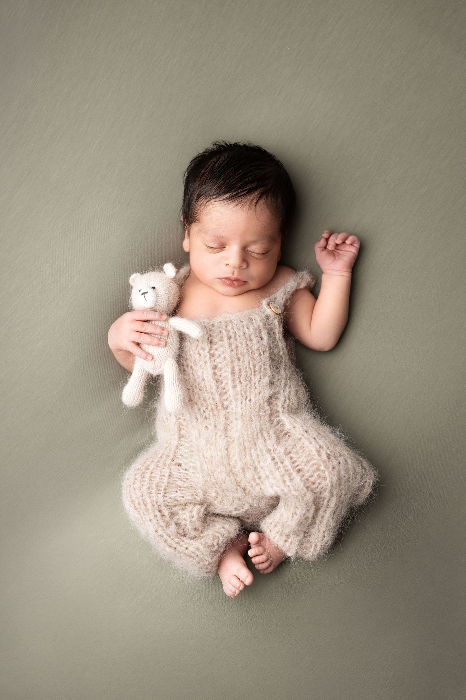 Sleeping newborn baby in knit romper holding tiny white teddy bear on sage green background.