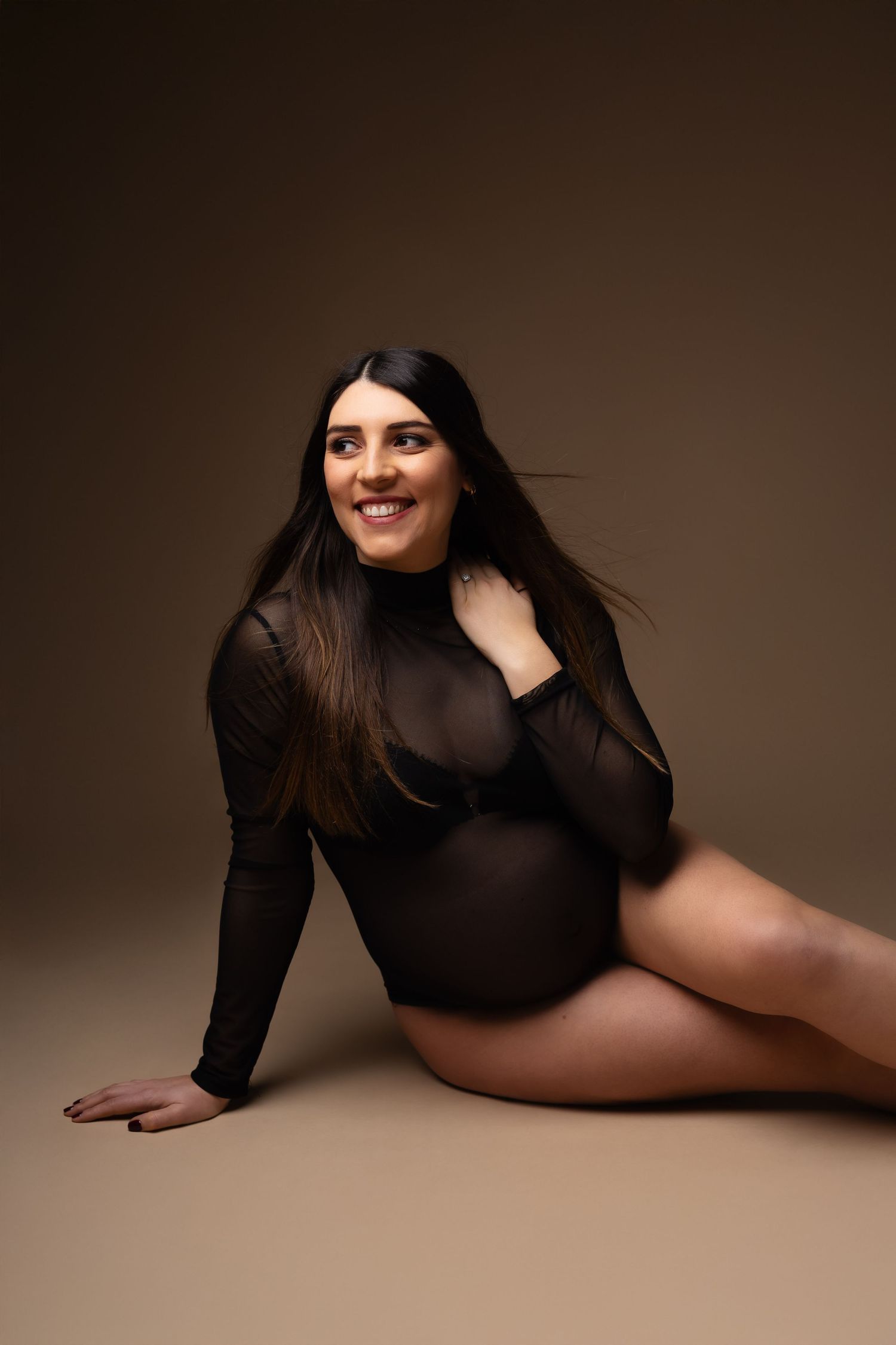 Happy pregnant woman in sheer black bodysuit sitting on floor, smiling, against warm brown studio background.