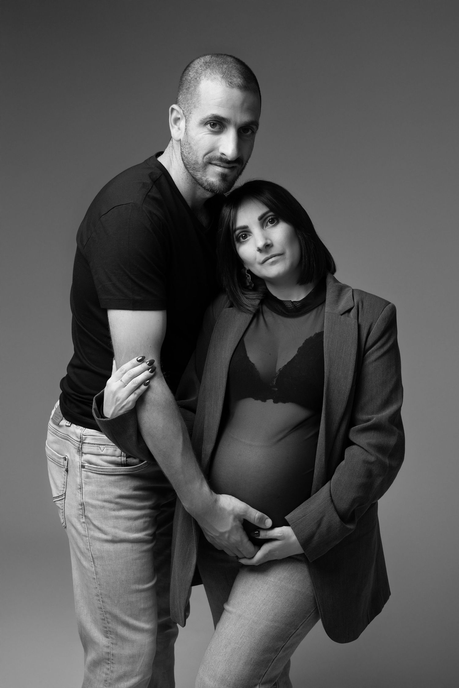Black and white studio photo of pregnant brunette woman embraced from behind by her partner. photographe grossesse chavanoz lyon