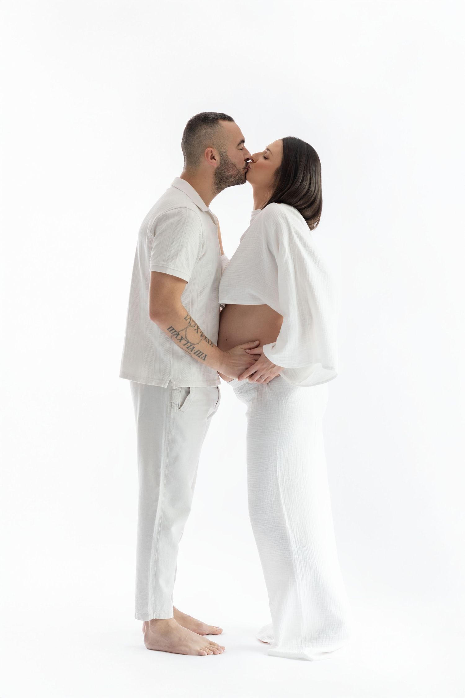 Pregnant couple kissing in matching all-white outfits on clean white studio background.