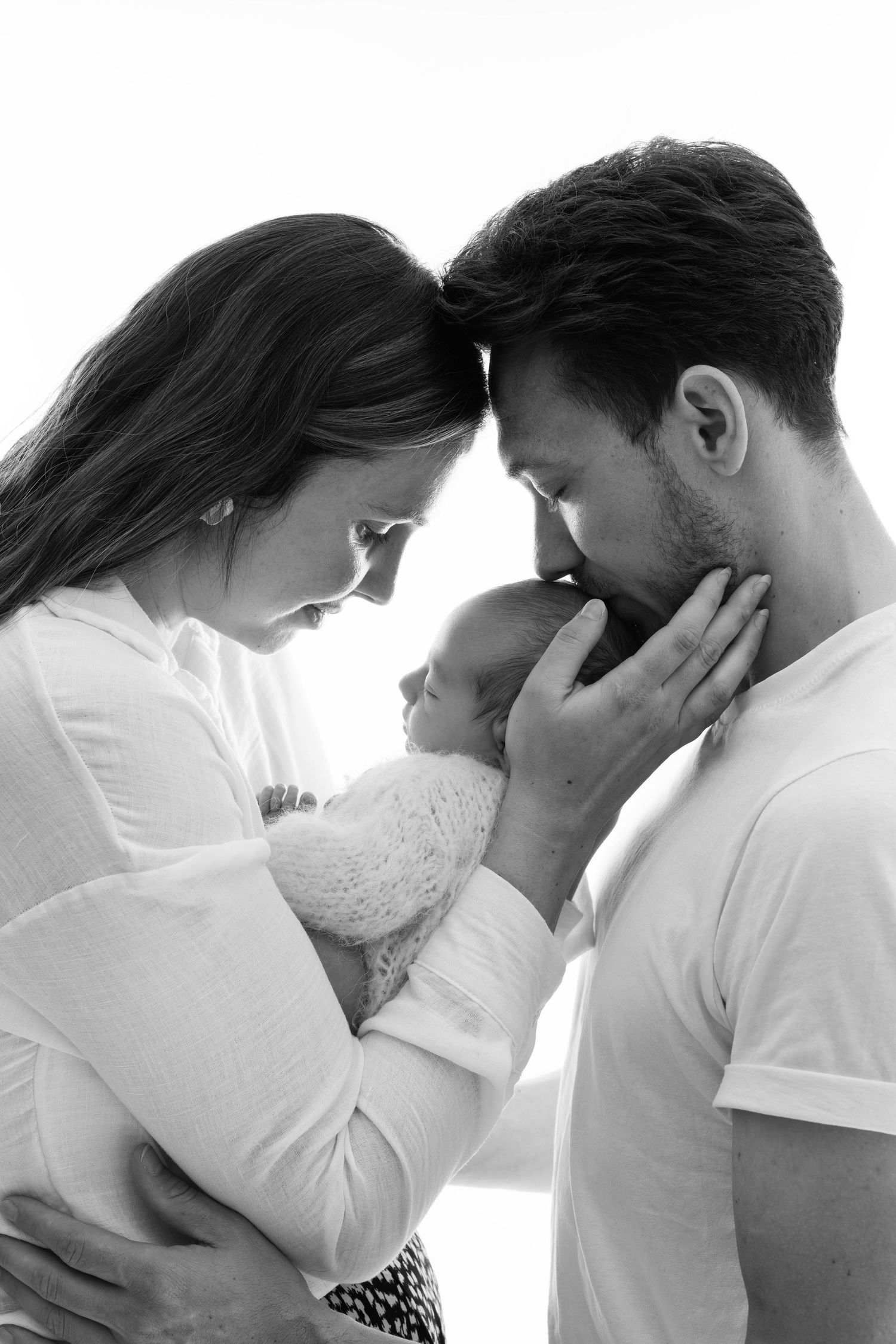 Black and white photo of parents tenderly holding their newborn baby, foreheads touching in an intimate family portrait.