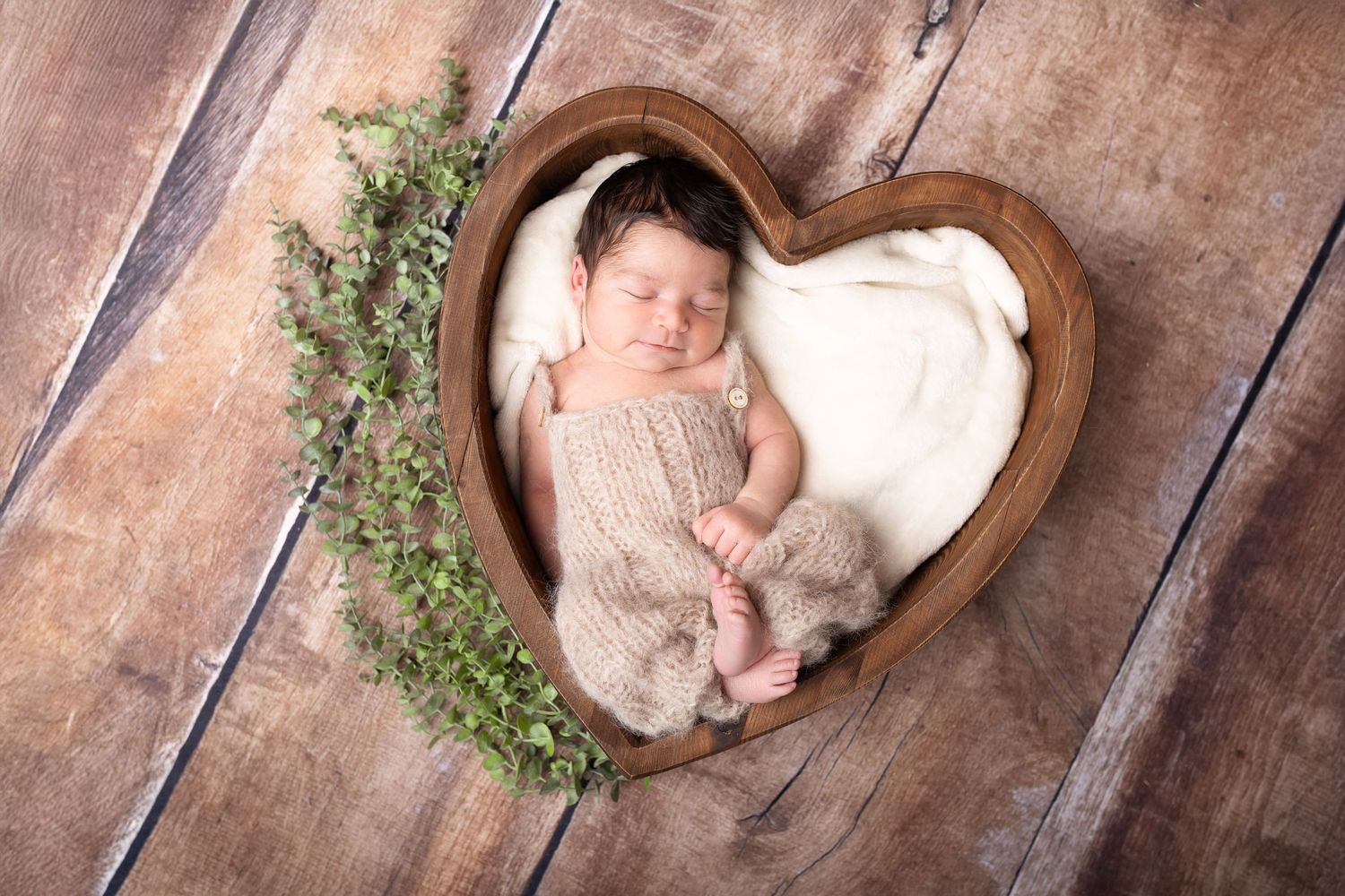 Sleeping newborn baby in knit romper posed inside a wooden heart-shaped bowl with green foliage decor. photographe naissance chavanoz lyon