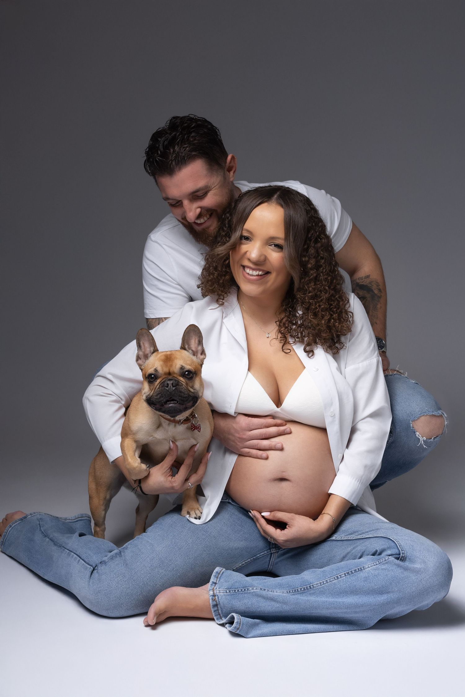 Smiling pregnant couple in white and jeans posing with French Bulldog in studio portrait.