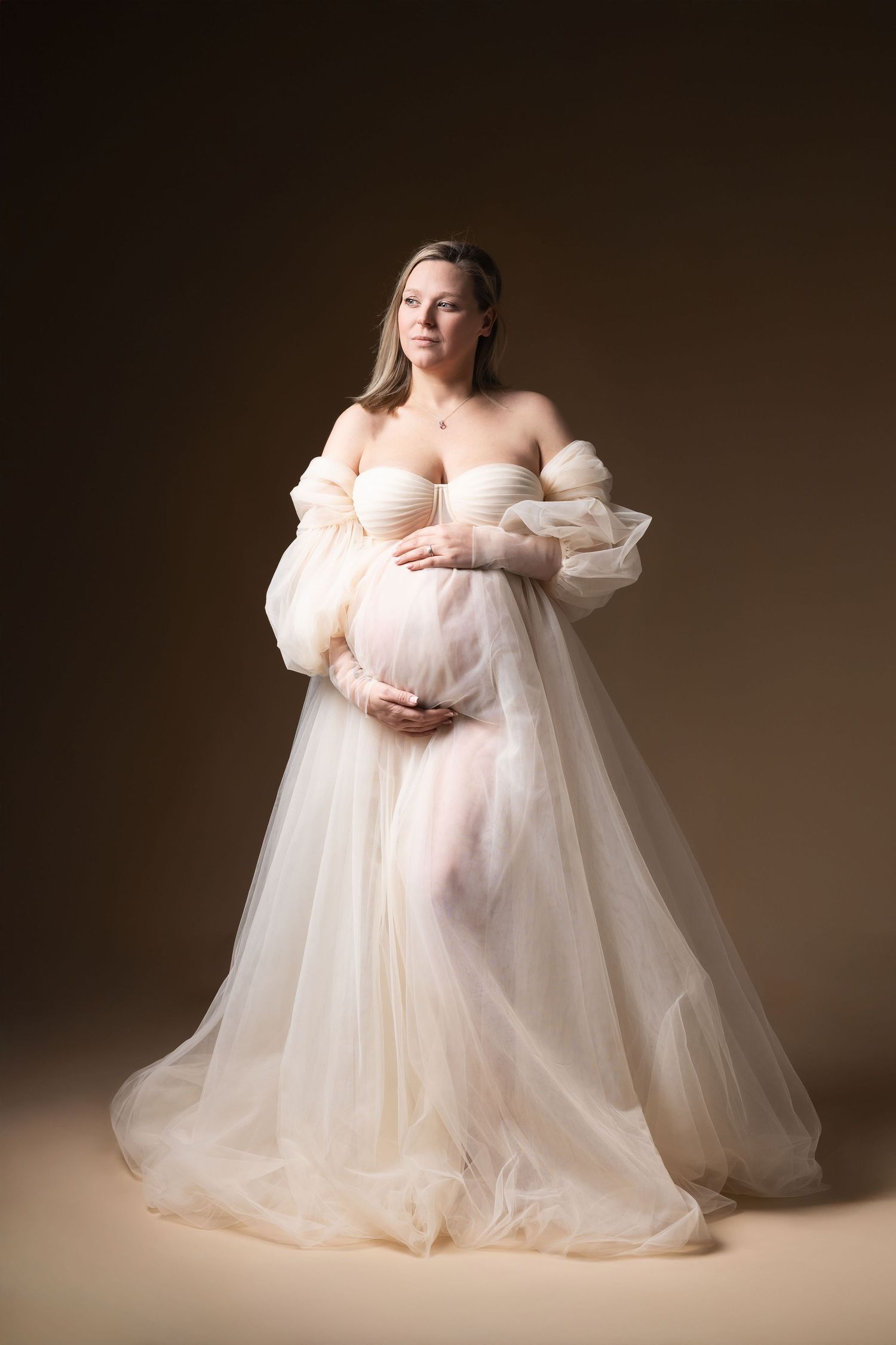 Pregnant woman in elegant cream tulle off-shoulder gown poses gracefully in studio portrait.