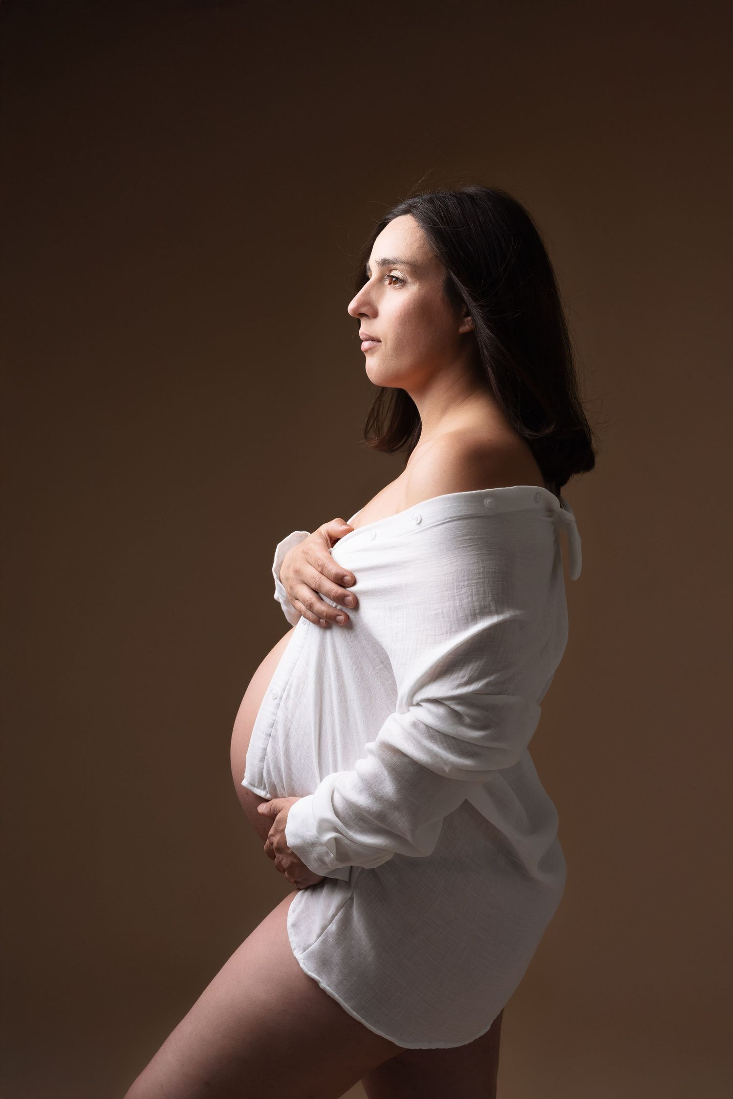 Elegant pregnant woman in white off-shoulder shirt cradling baby bump on brown background. photographe grossesse chavanoz lyon