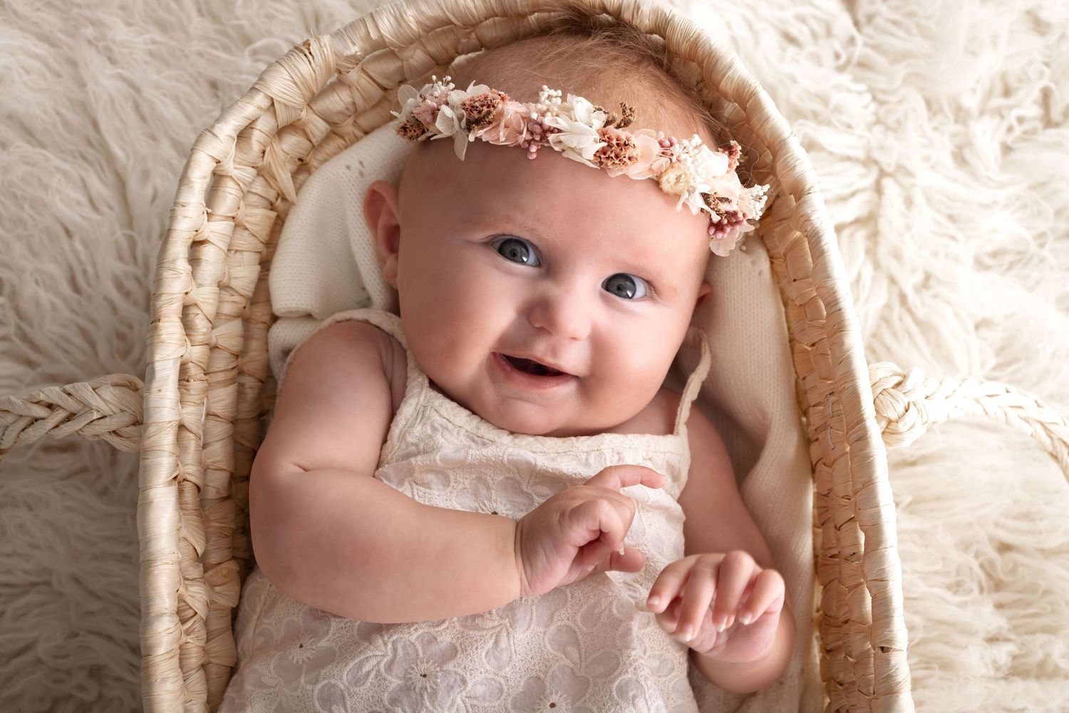 Smiling baby girl in white lace dress and floral headband lying in wicker basket on fluffy white fur background.
