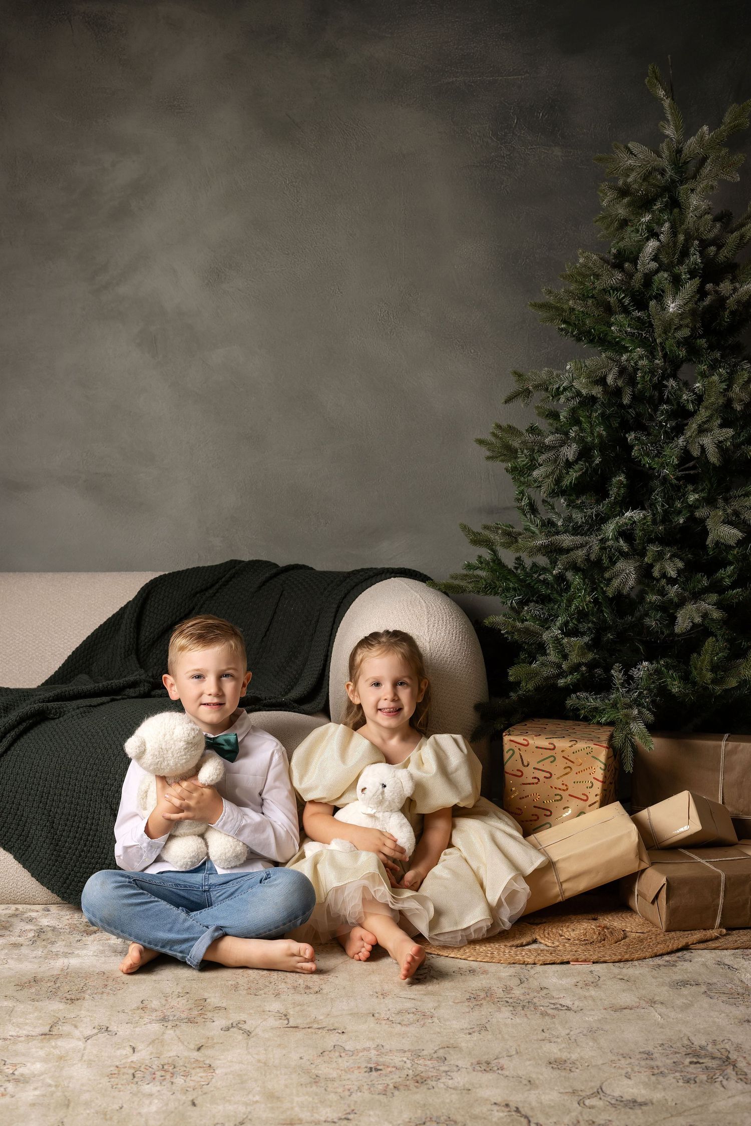 Two children sit barefoot by a Christmas tree holding stuffed animals, surrounded by wrapped gifts in a cozy studio setting.