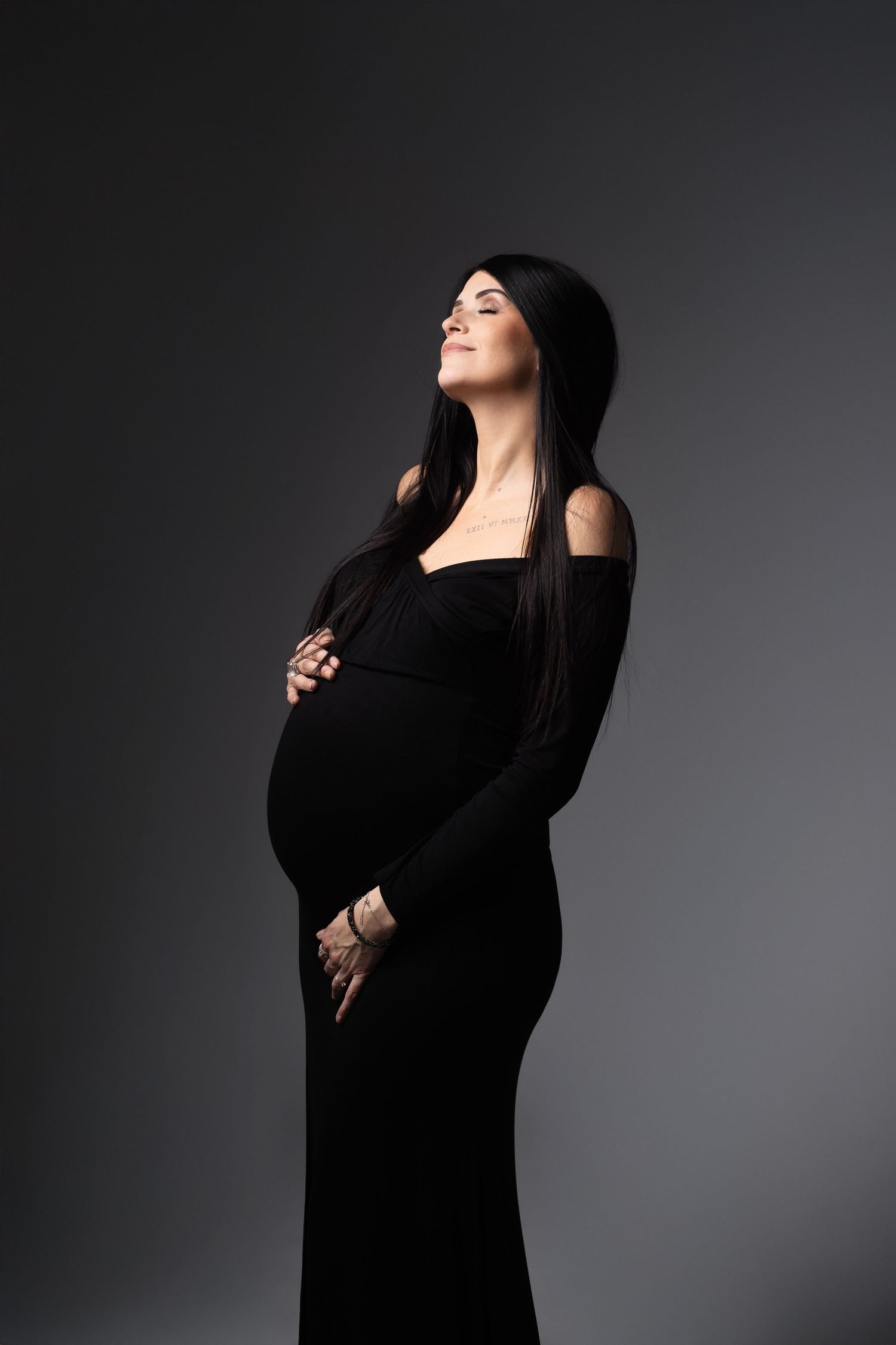 Peaceful pregnant woman in black dress holding baby bump with eyes closed against dark gray background.