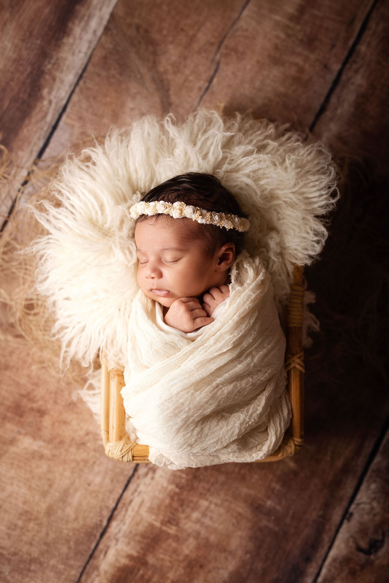 Sleeping newborn baby wrapped in cream swaddle with floral headband resting in a small wooden basket with fluffy white fur.