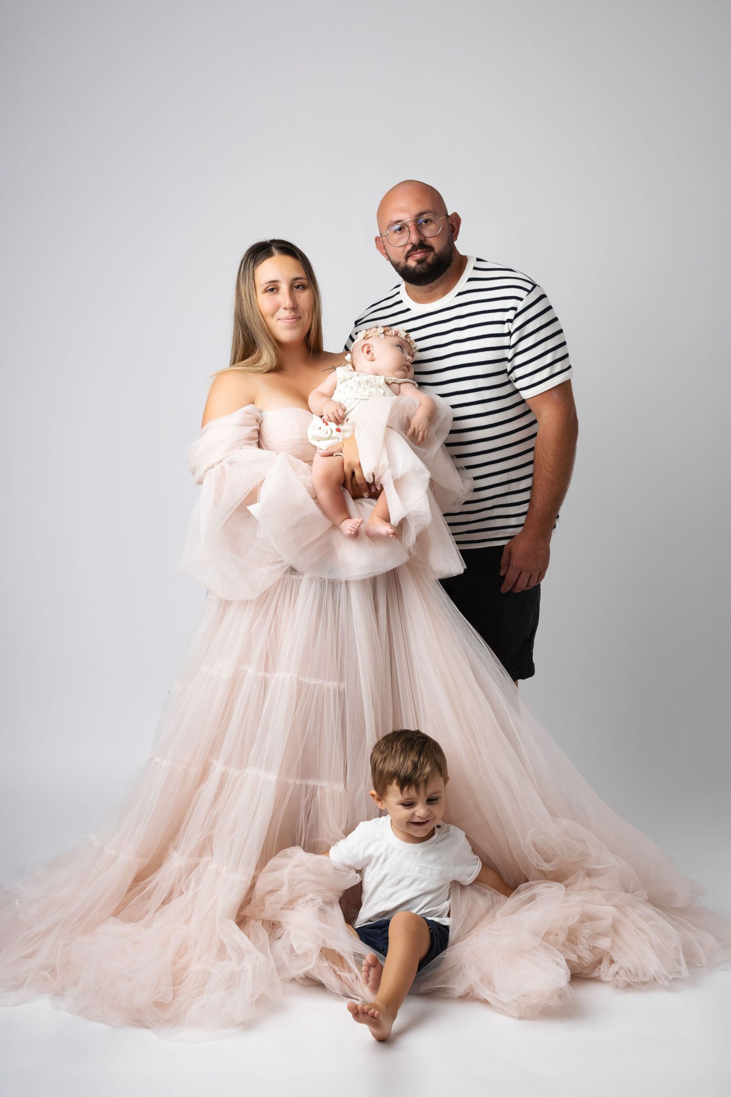 Family portrait with mother in pink tulle gown holding baby, father in striped shirt, and young boy sitting on floor.