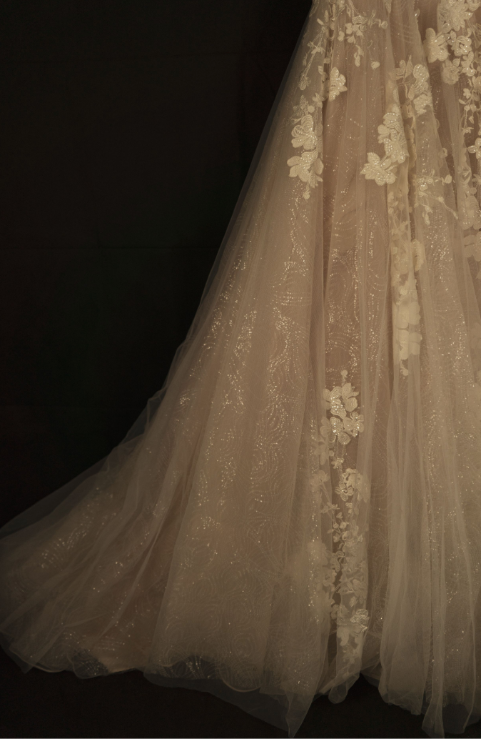 Close-up of a champagne coloured wedding dress train featuring delicate white floral lace appliques against a dark background.