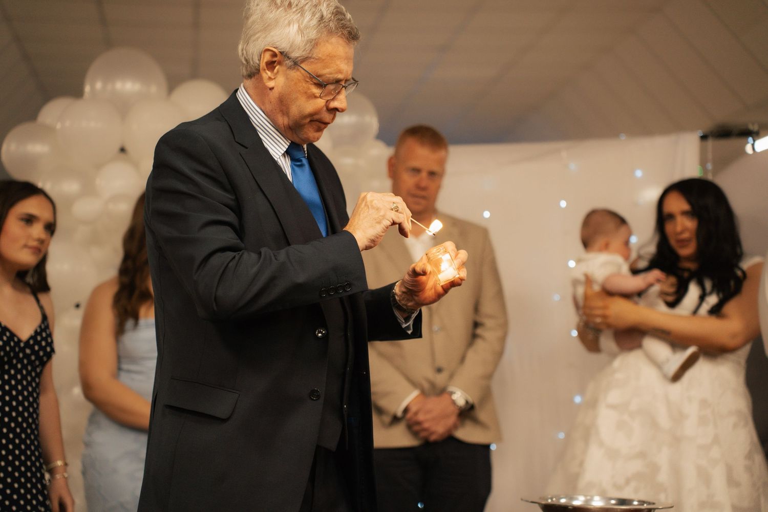 Celebrant in a dark suit lighting a candle while a family, including a woman holding a baby, watches in a room decorated with white balloons