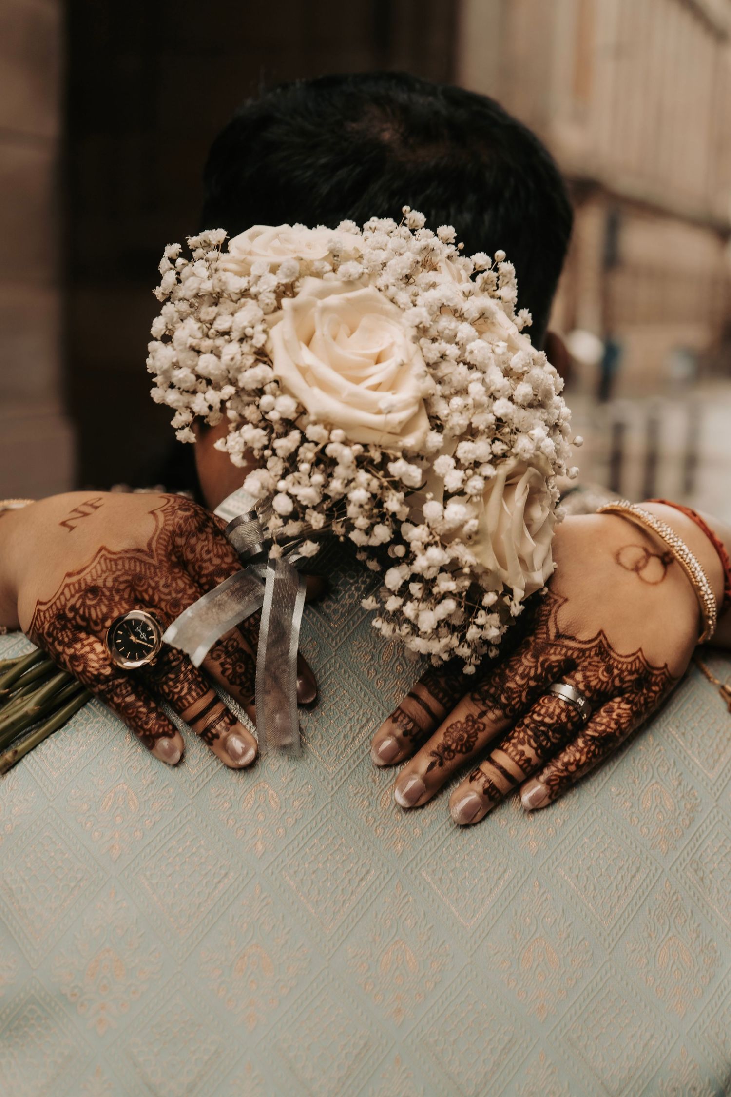 Bride’s henna adorned hands holding a bouquet of white roses and baby’s breath while embracing her partner in light green attire