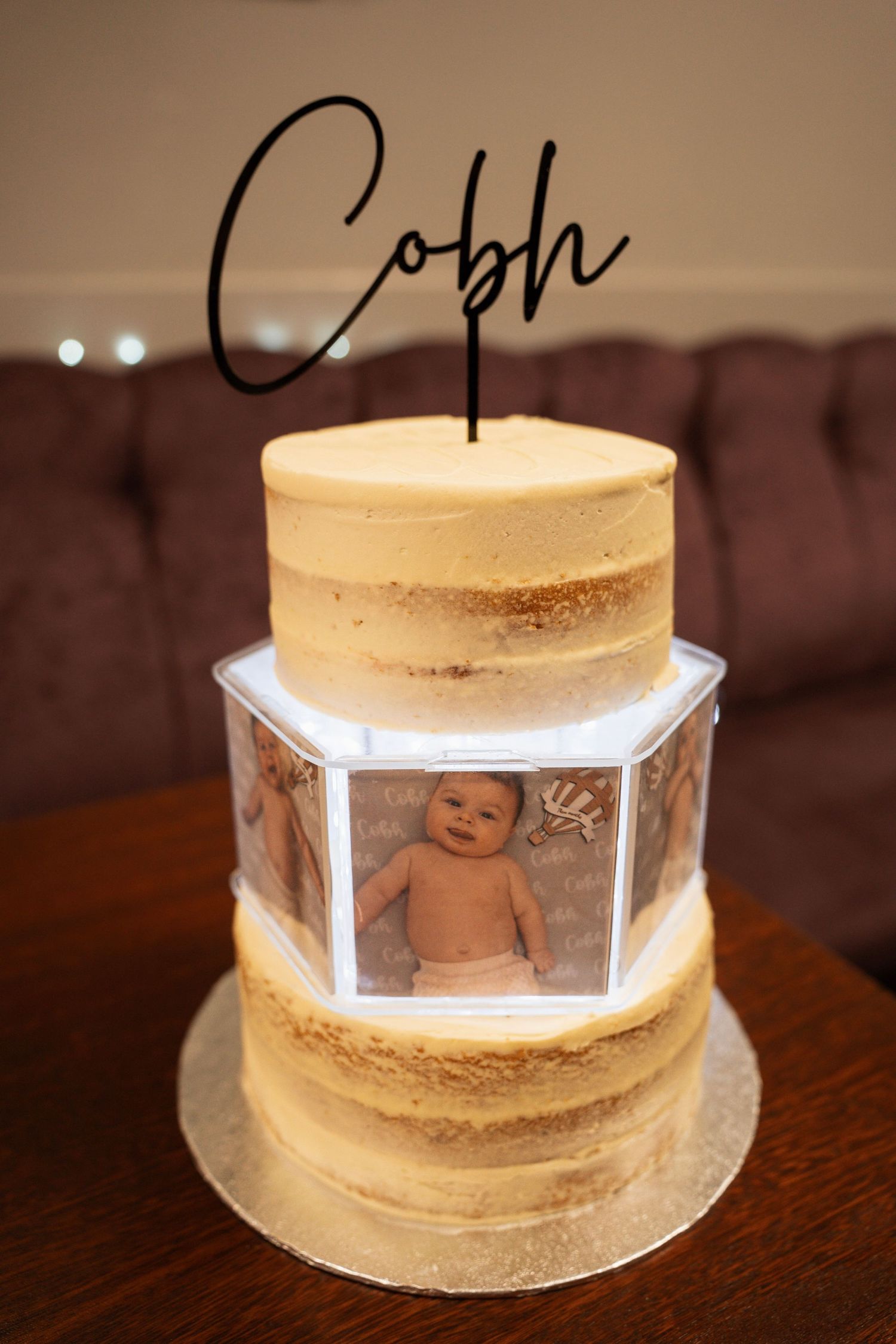Two-tiered naked cake featuring a photo display of a baby in the center tier and a black script cake topper