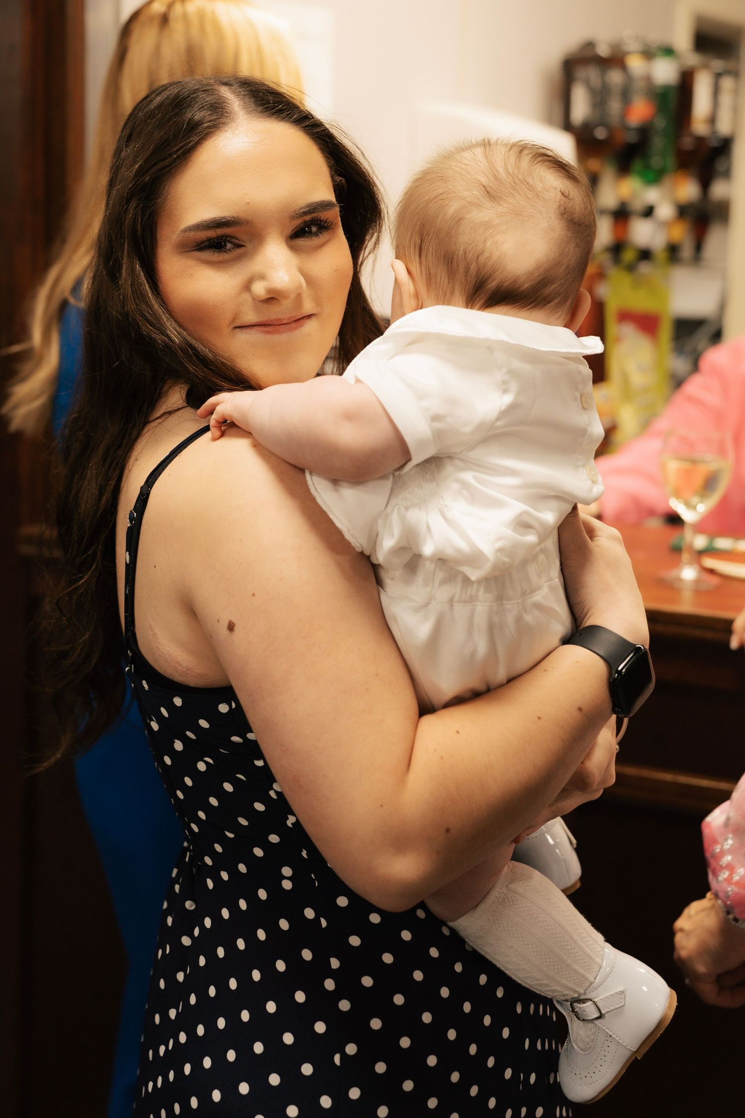 Young woman in a black polka-dot dress smiles while holding a baby dressed in a white outfit at a christening.