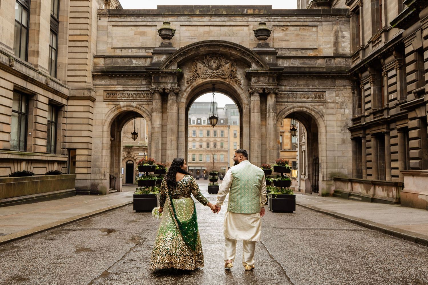 Bride and groom in green and gold traditional attire, walking hand in hand under the arches in Glasgow, in a candid city wedding photo
