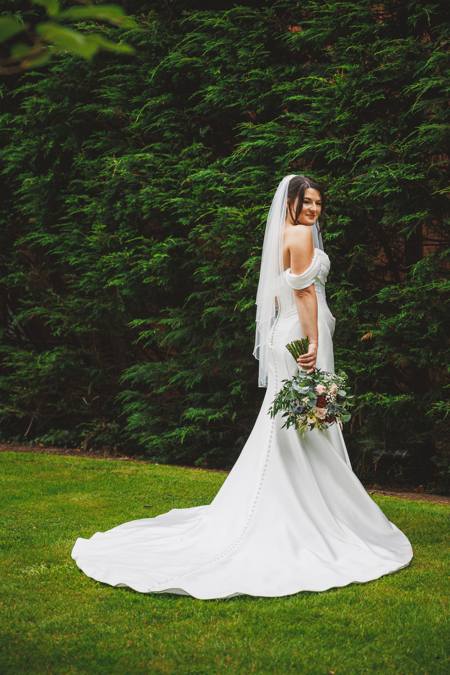Bride in backless white gown with veil holds bouquet on green lawn before lush hedge backdrop.