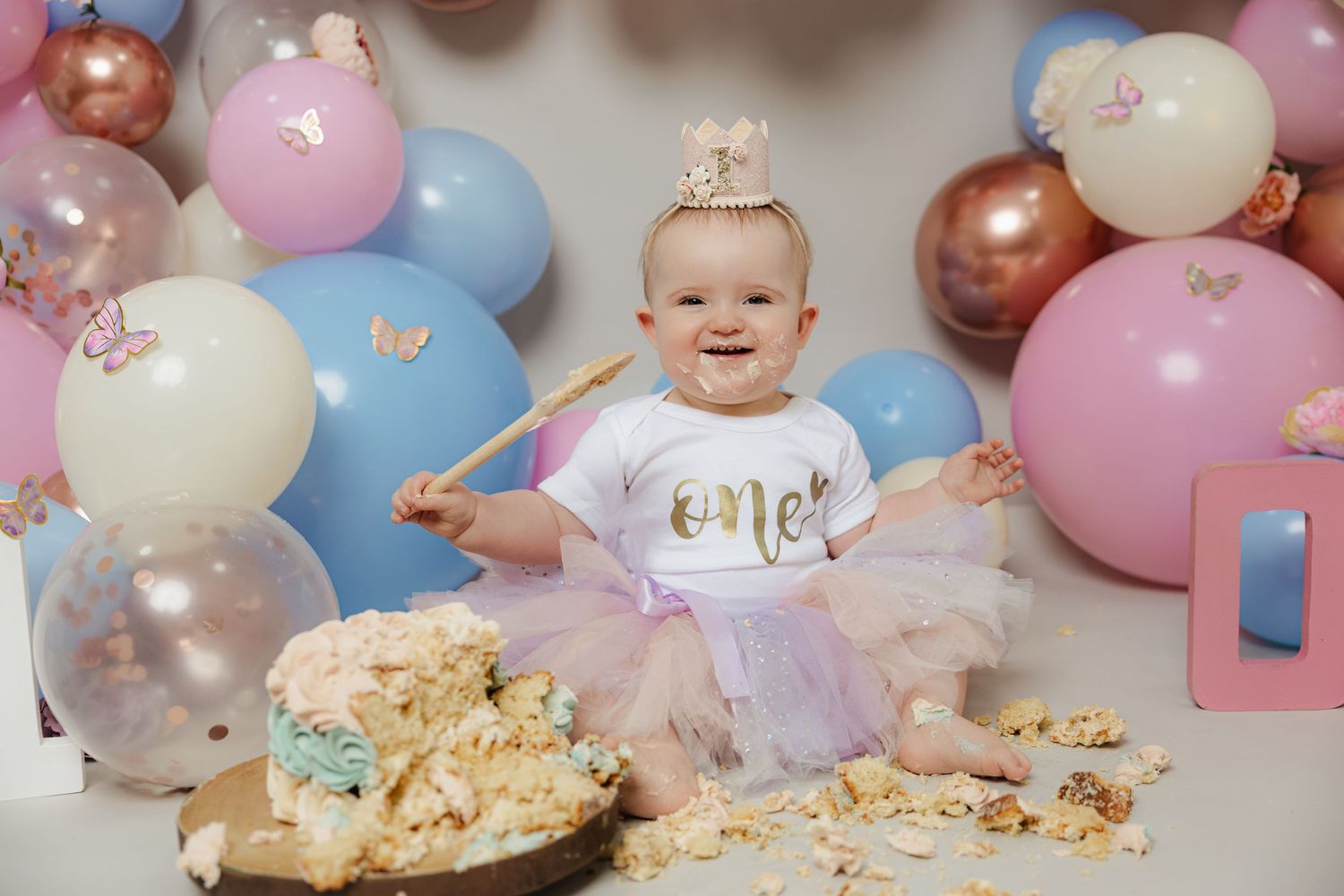 Baby girl in tutu celebrates first birthday smashing cake surrounded by pink and blue balloons.