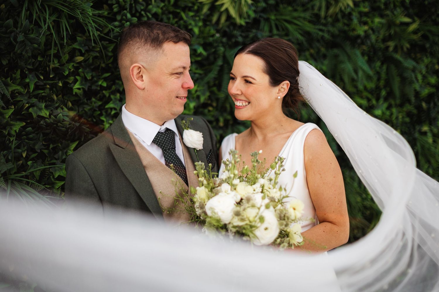 Smiling bride holding white bouquet stands with groom in gray suit before lush green hedge on wedding day.