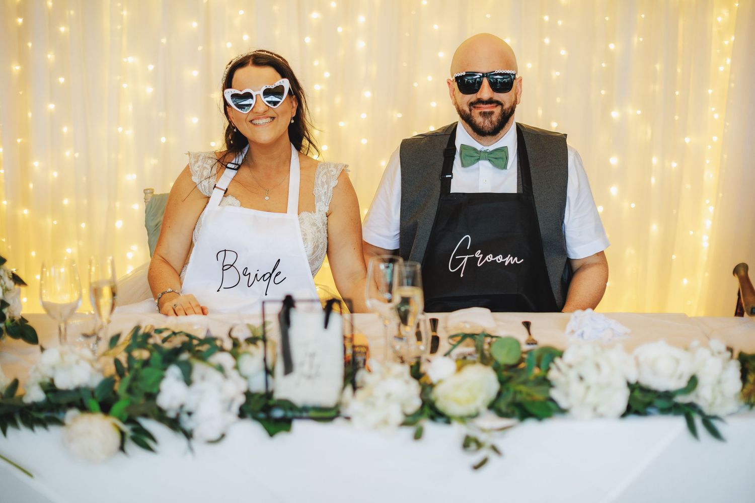 Bride and groom wearing aprons and novelty sunglasses pose at floral-decorated wedding reception head table.