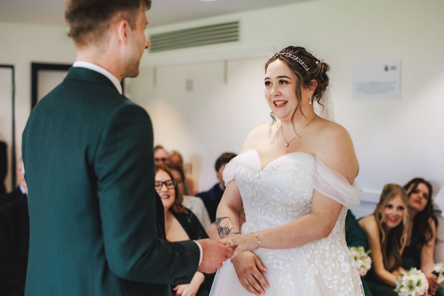 Bride in off-shoulder lace gown smiles joyfully at groom during wedding ceremony, guests seated in background.