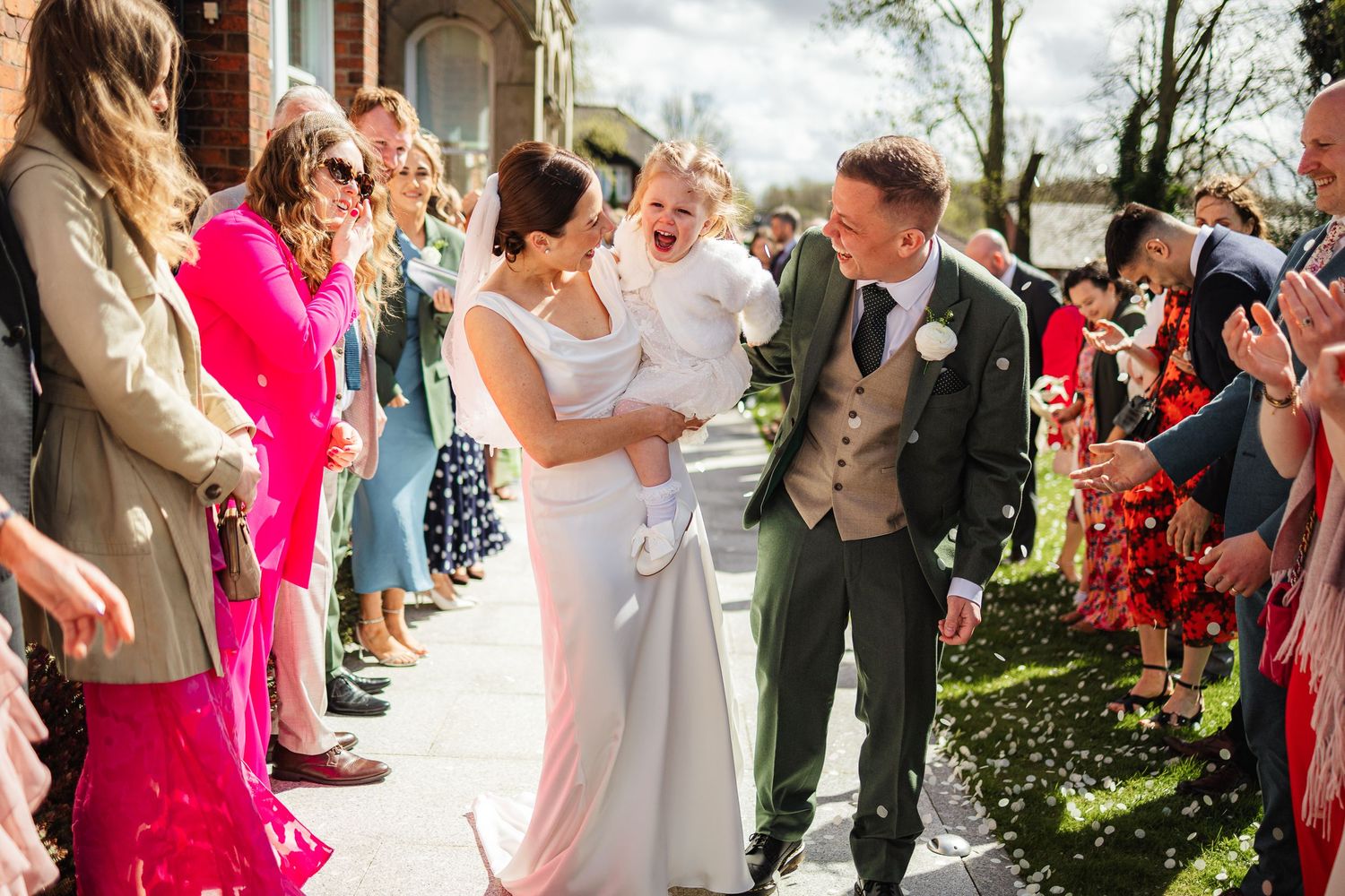 Bride and groom walk through confetti with baby at outdoor wedding celebration surrounded by smiling guests.