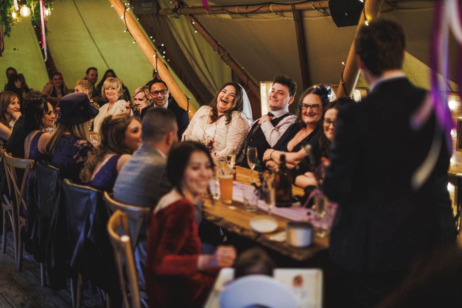 Wedding guests laughing and celebrating at a rustic tipi reception dinner, bride and groom seated at long wooden table.