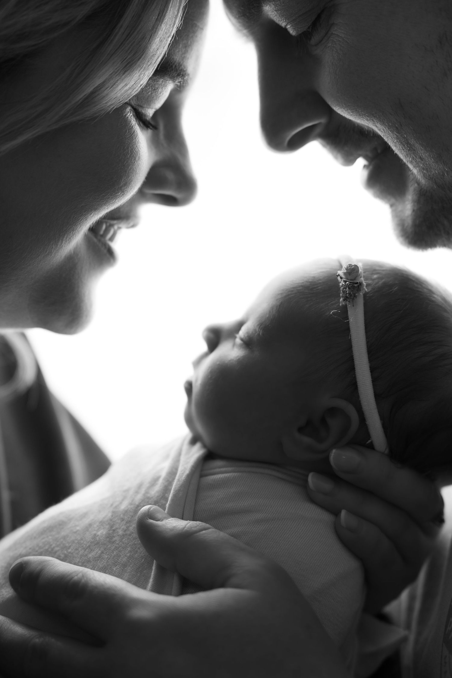 Black and white photo of smiling parents tenderly holding their sleeping newborn baby girl wearing a white headband.