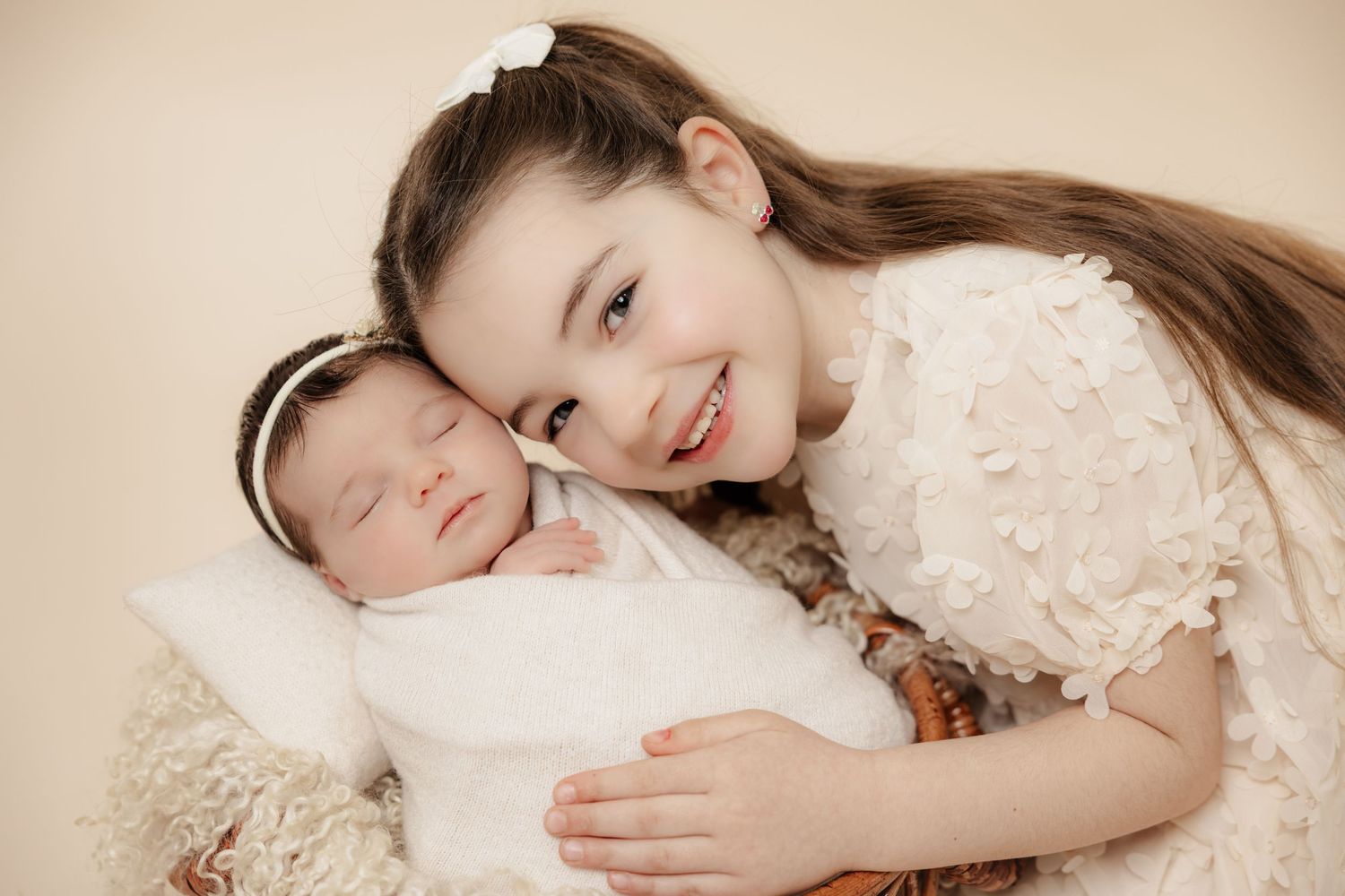 Young girl in white dress smiles beside sleeping newborn baby in a soft, warm-toned portrait photo.