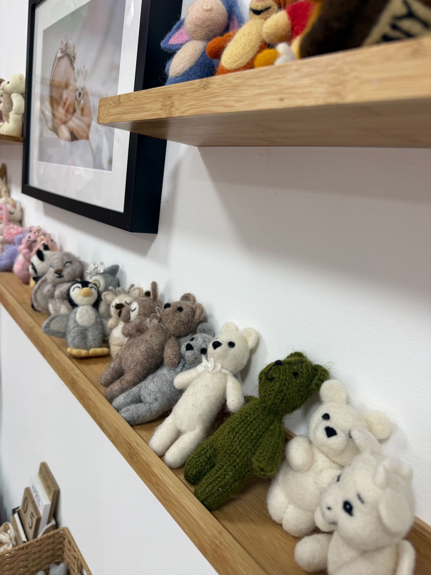 Wooden shelves displaying a collection of handmade felted and knitted teddy bears and animals, with a baby photo frame on the wall.
