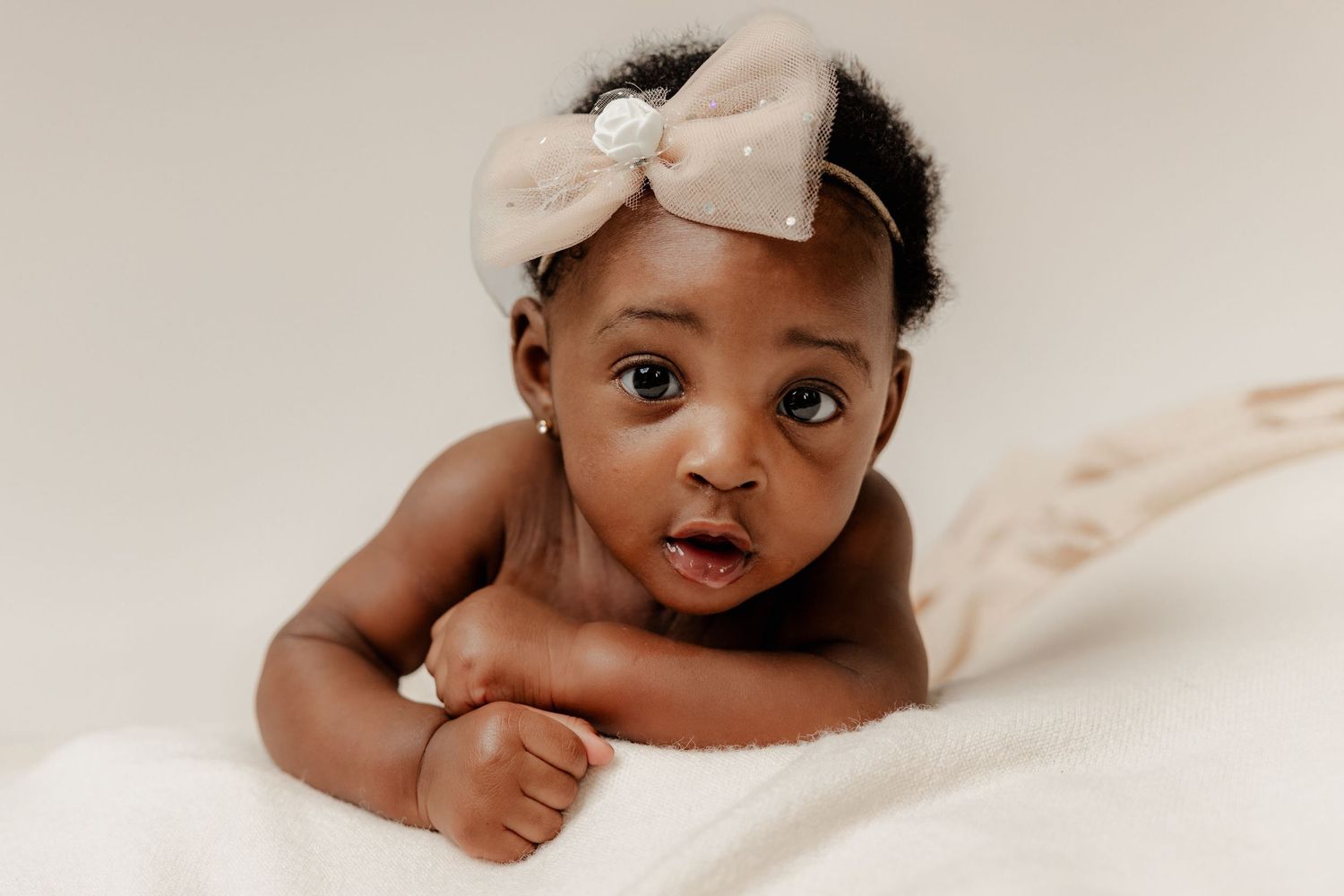 Adorable dark-skinned baby girl wearing a beige bow headband lying on a soft white blanket looking at camera.