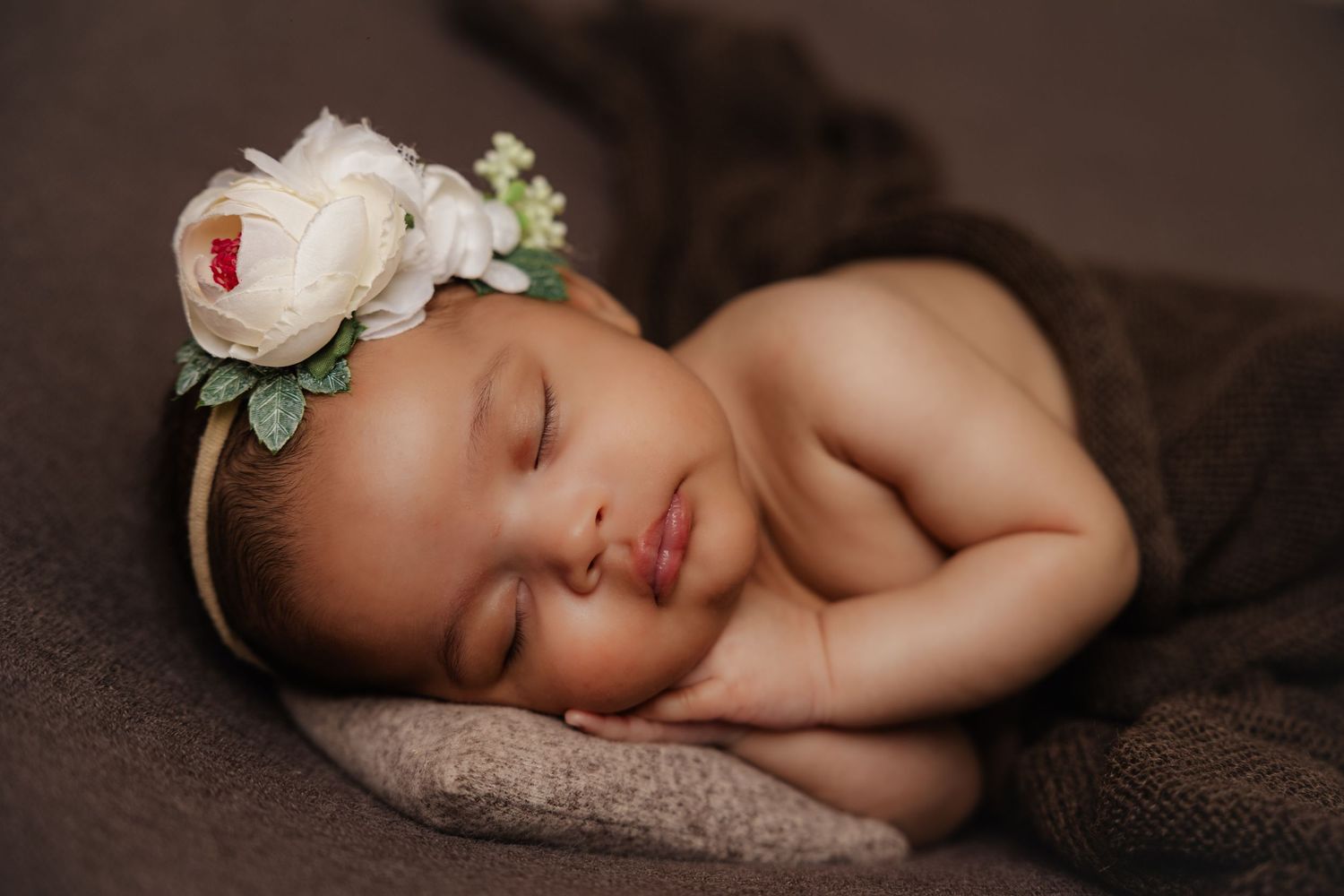 Peaceful newborn baby girl sleeping with white floral headband, wrapped in brown blanket in professional portrait.