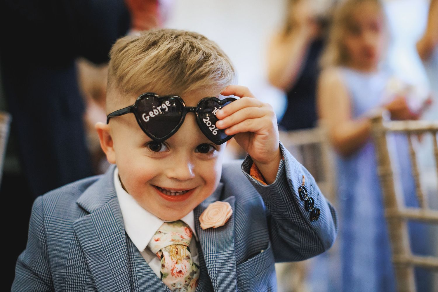 A smiling boy in a blue suit holds up heart-shaped sunglasses at a wedding celebration.