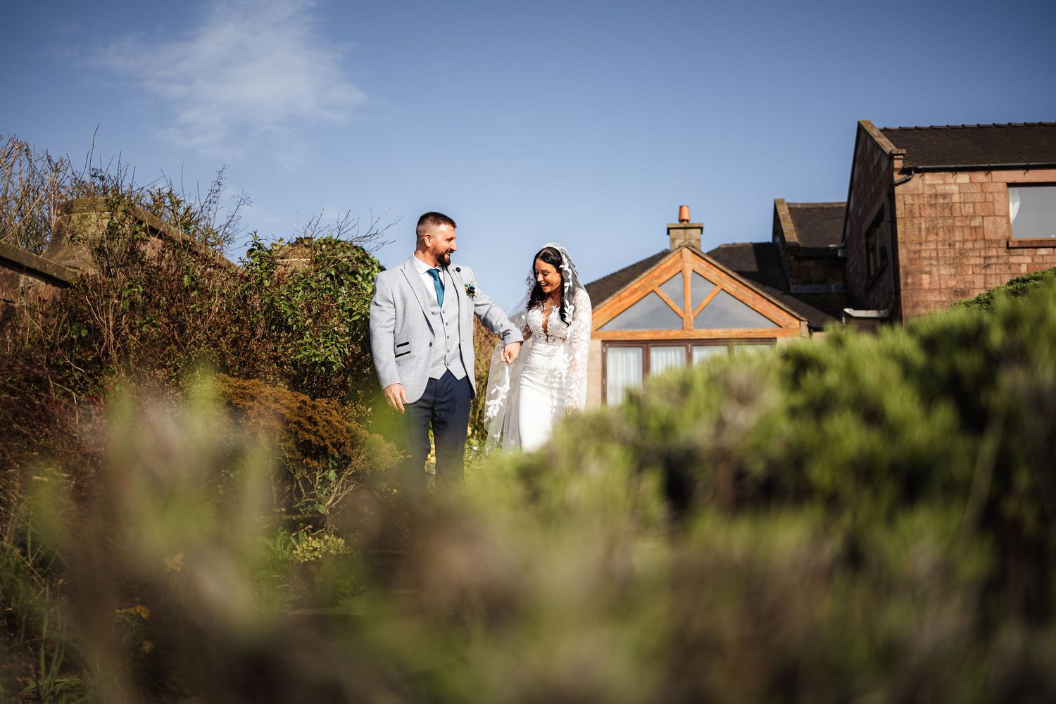 A bride and groom stand together in a lush garden outside a brick venue under a bright blue sky.