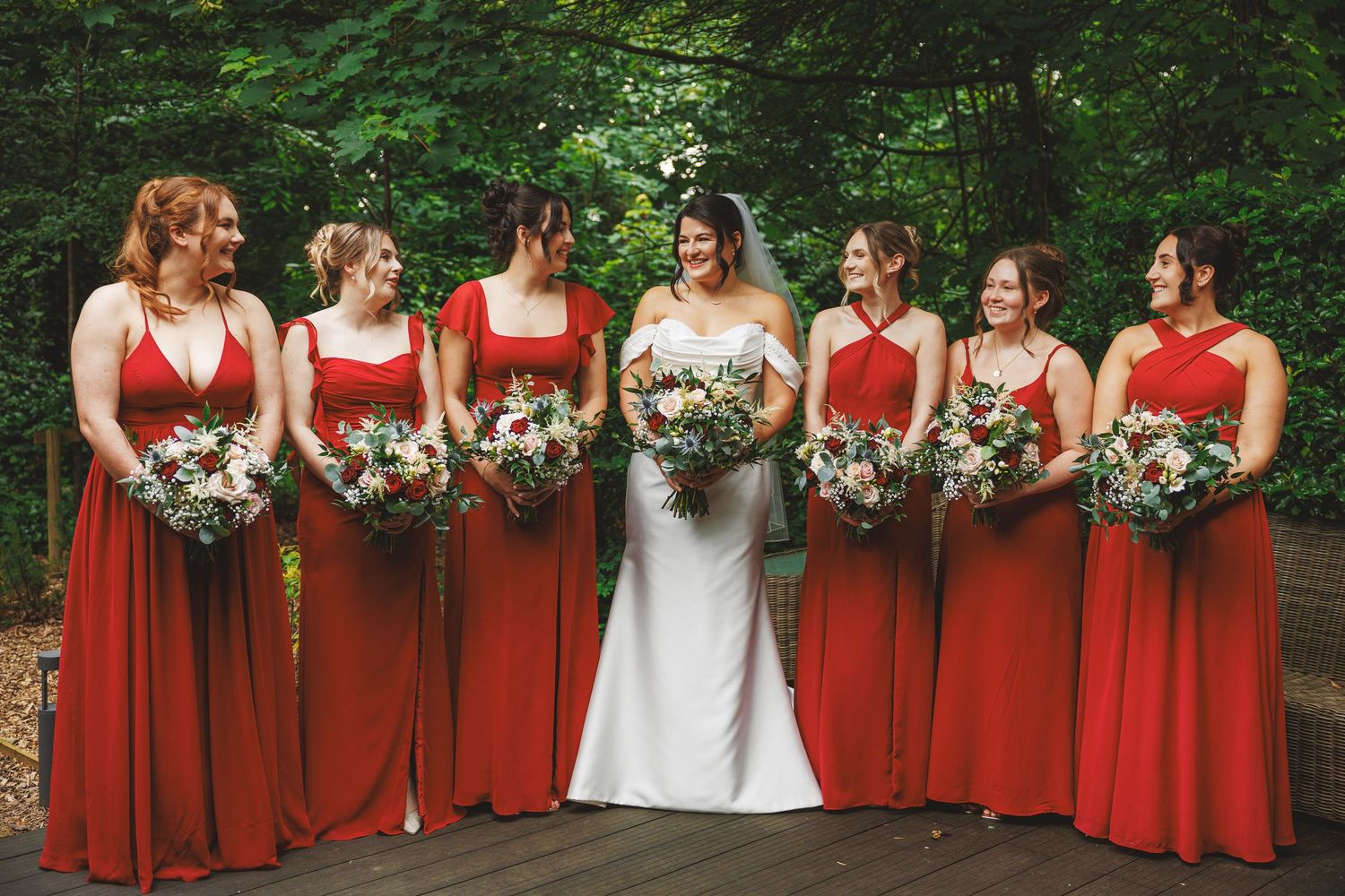 Bride in white gown stands with six bridesmaids in red dresses holding floral bouquets on a wooden deck outdoors.